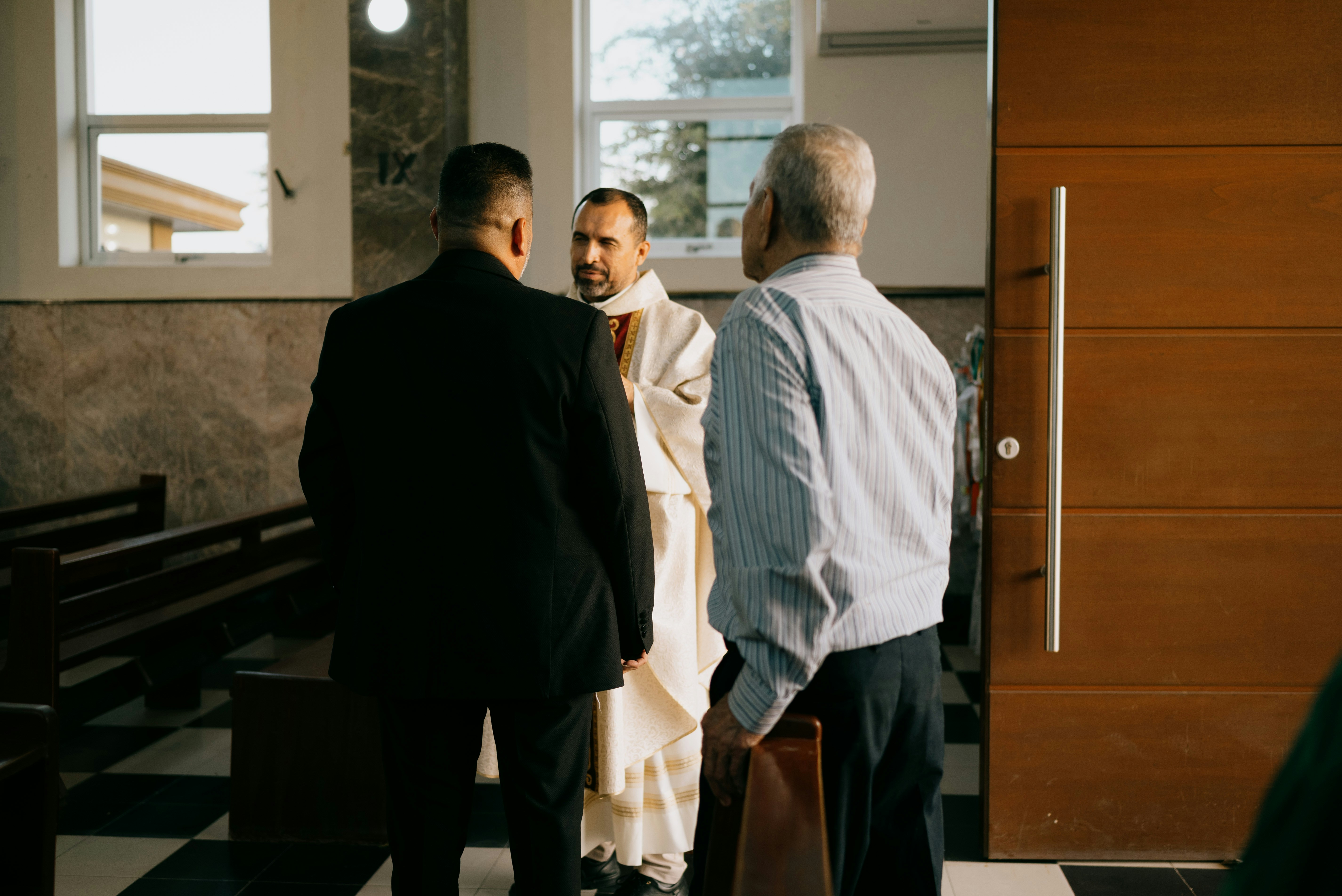 Cleric in white vestments speaks with two men.
