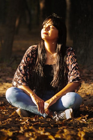 A woman meditates outdoors in a forest setting.