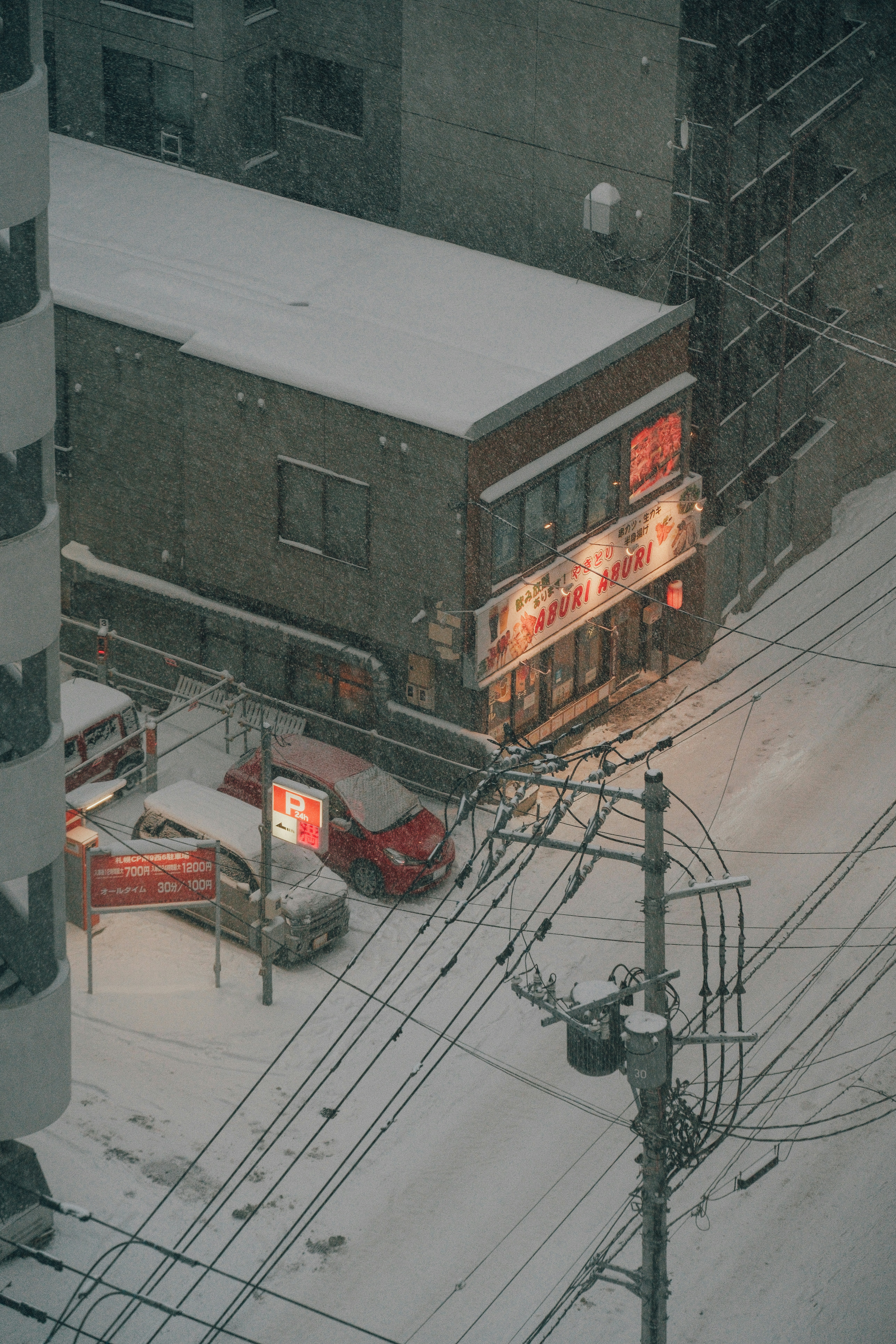 Snow falls on a city street with cars and buildings.