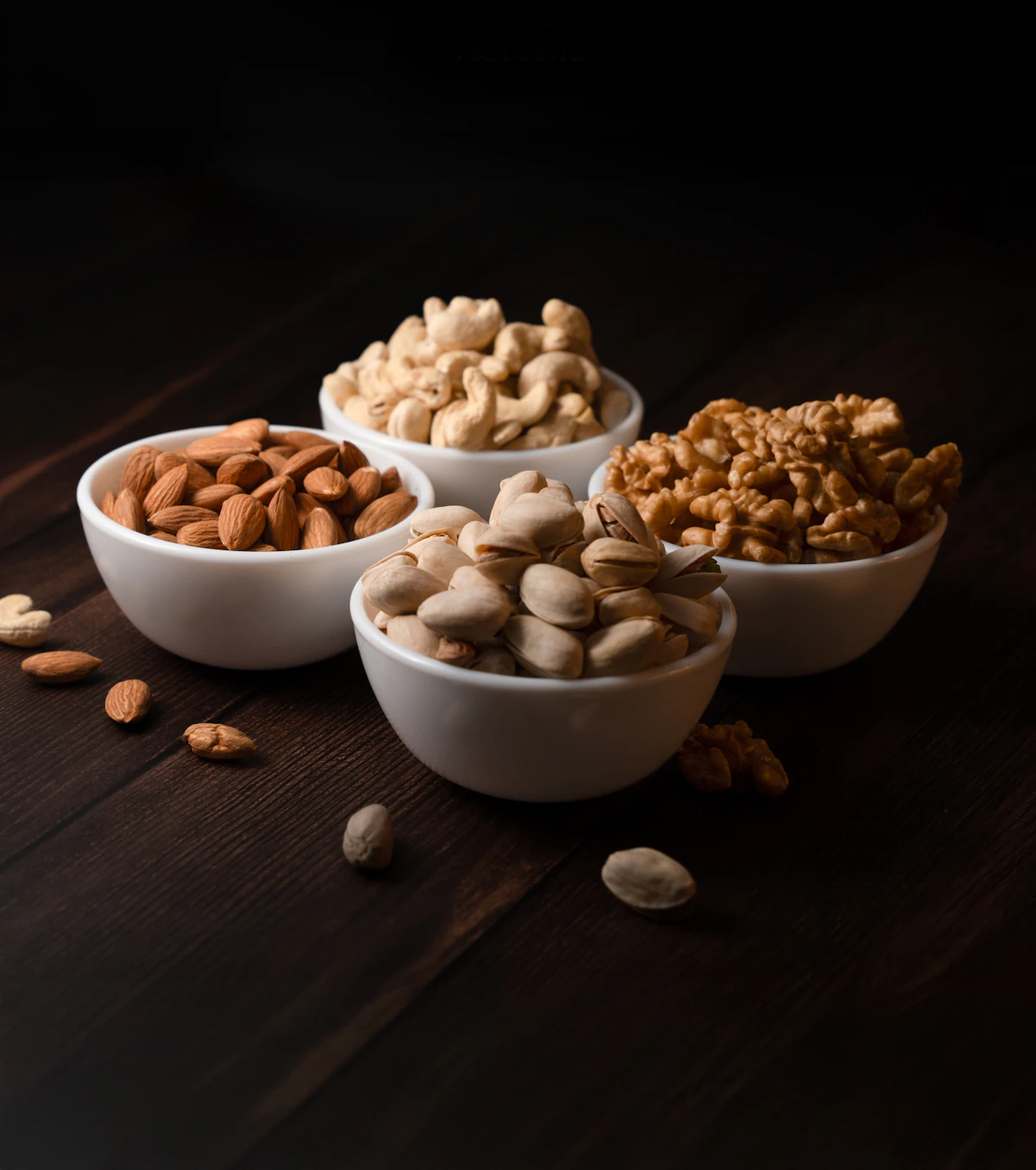 Almonds, cashews, pistachios, and walnuts set out in four open bowls on dark wood