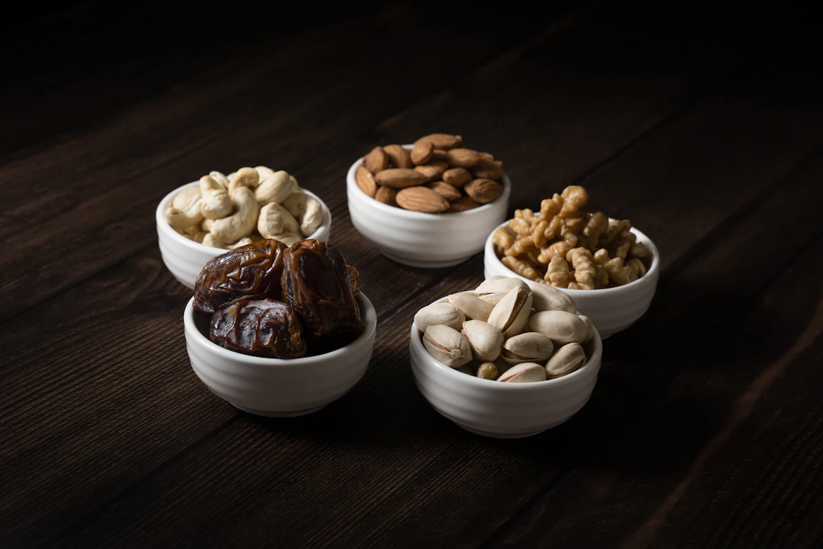 Cashews, almonds, walnuts, dates, and pistachios in five small bowls on dark wood