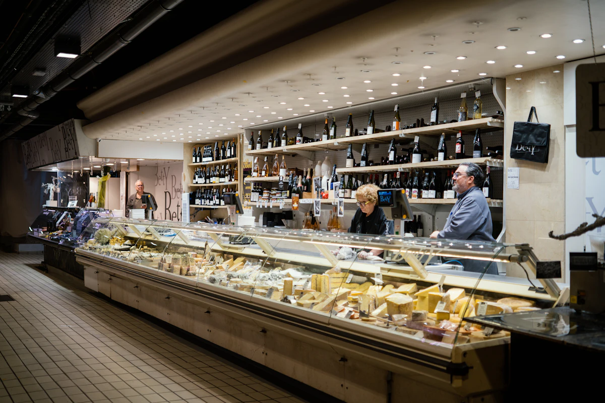Un long comptoir réfrigéré garni de fromages à la coupe dans une halle, avec deux vendeurs derrière la vitrine et des rayonnages de bouteilles au mur