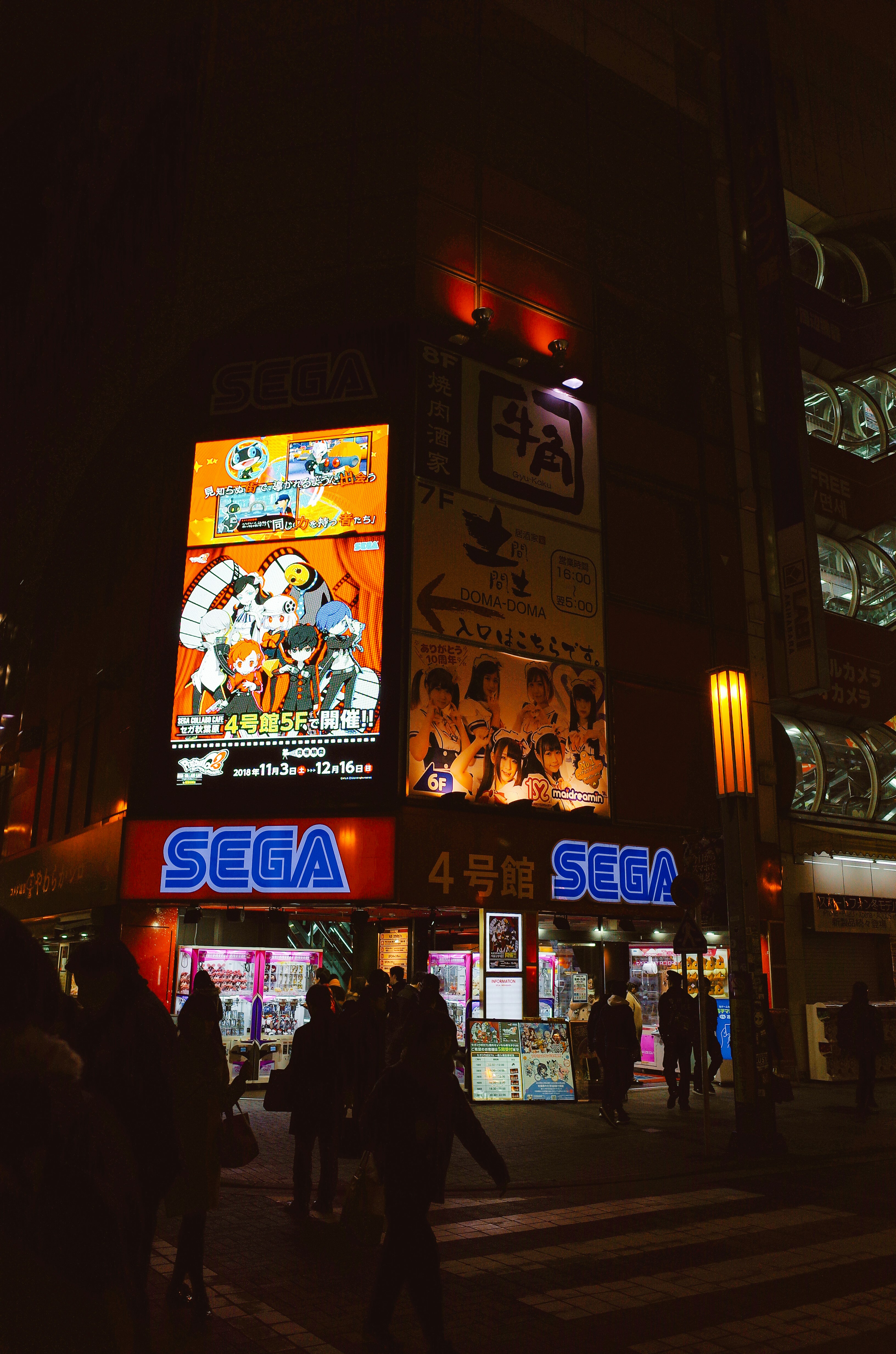 A brightly lit sega arcade at night with people walking.