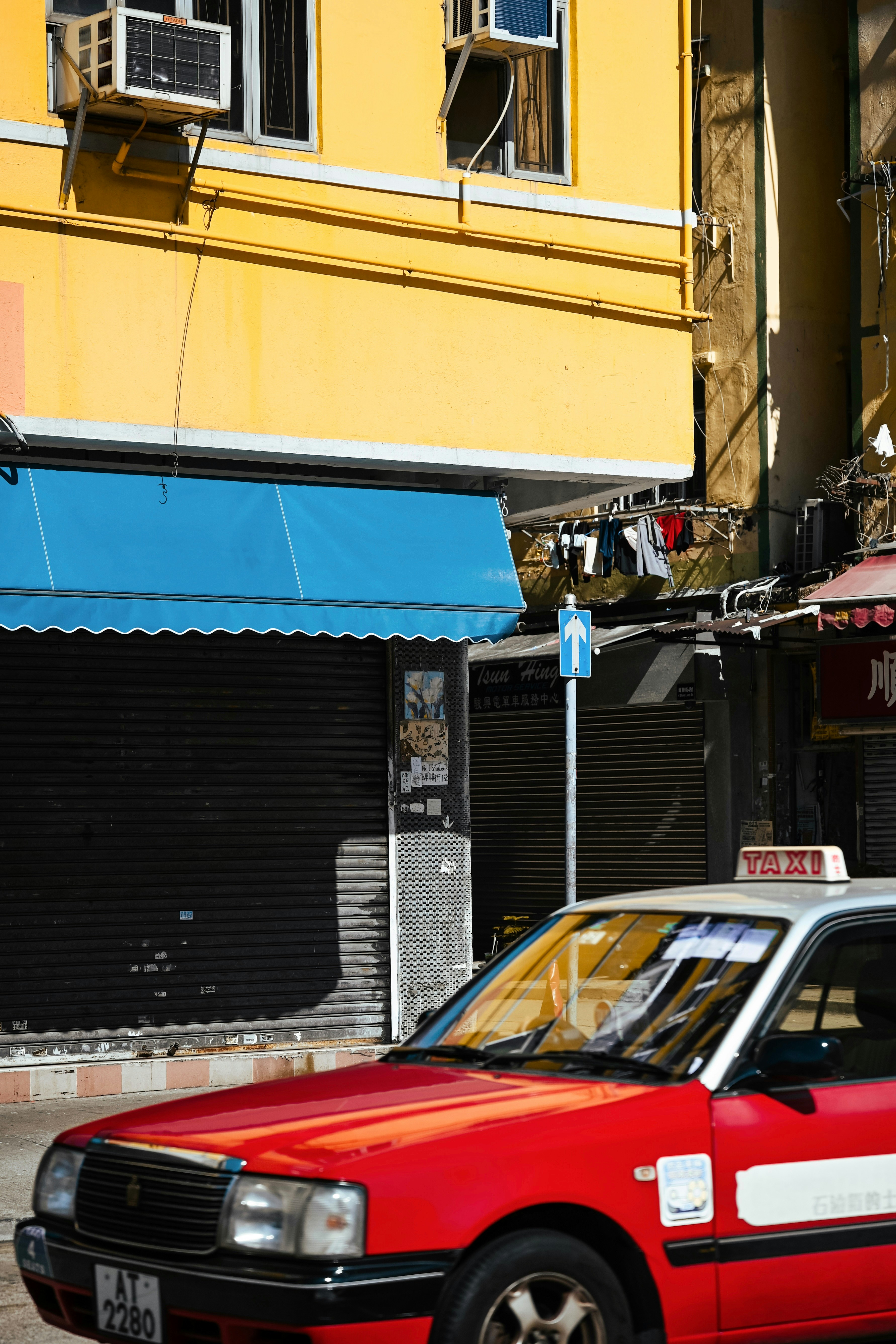 Red taxi drives past yellow building and blue awning.