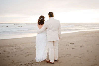 Newlyweds embrace on a sandy beach at sunset.