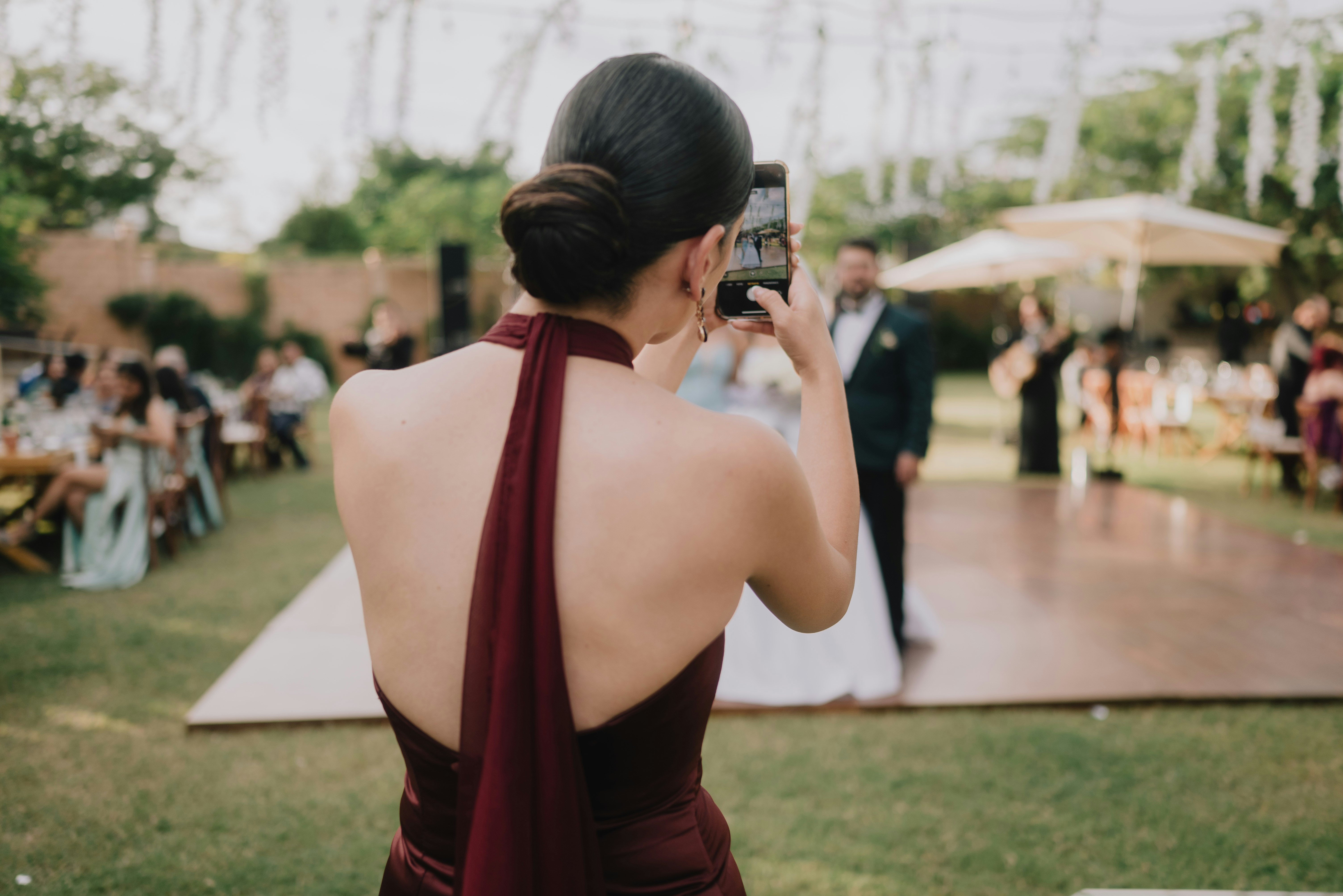 Une femme en robe bordeaux photographie un couple lors d’un événement en plein air, dans une ambiance festive.