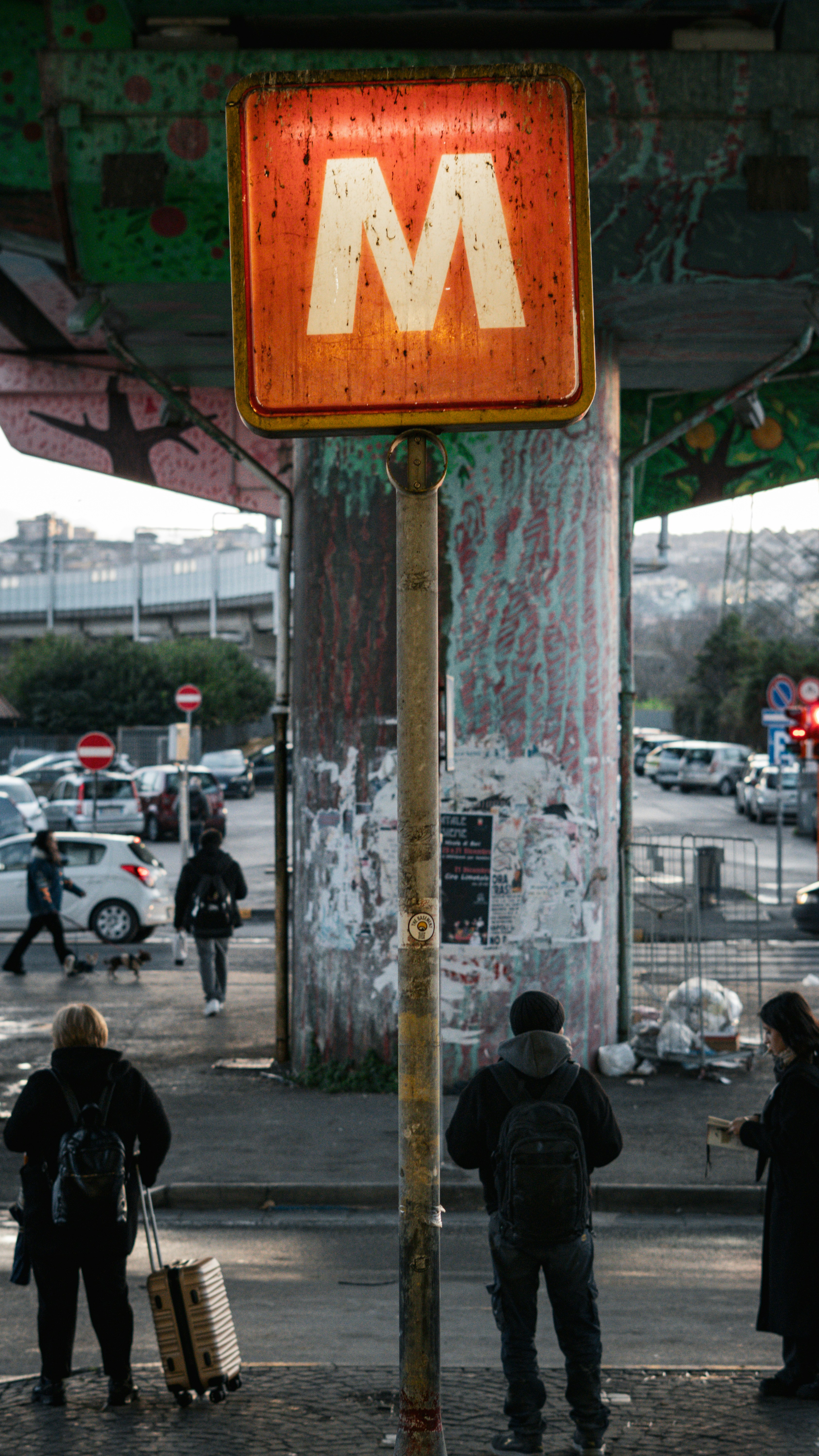 People wait near a weathered metro sign in a city.
