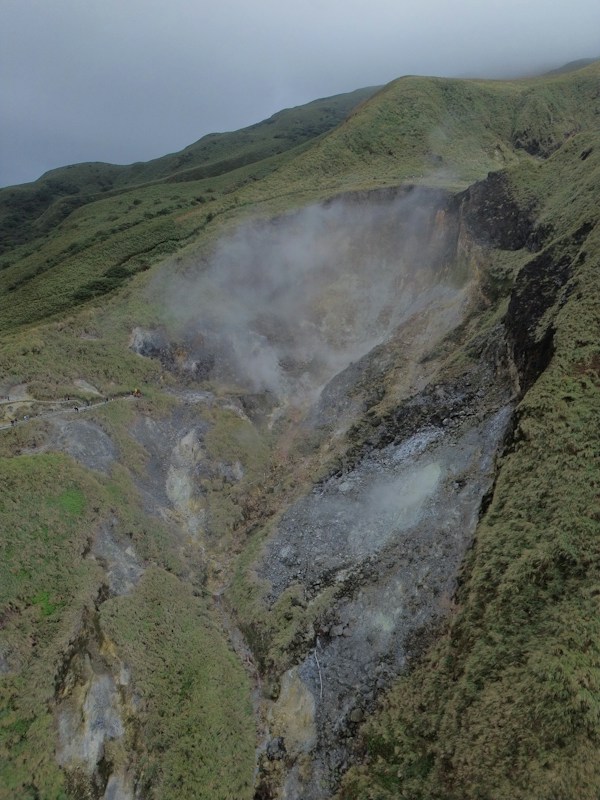 台灣山區草坡上的火山噴氣孔冒出白色蒸氣，左側有步道與遊客