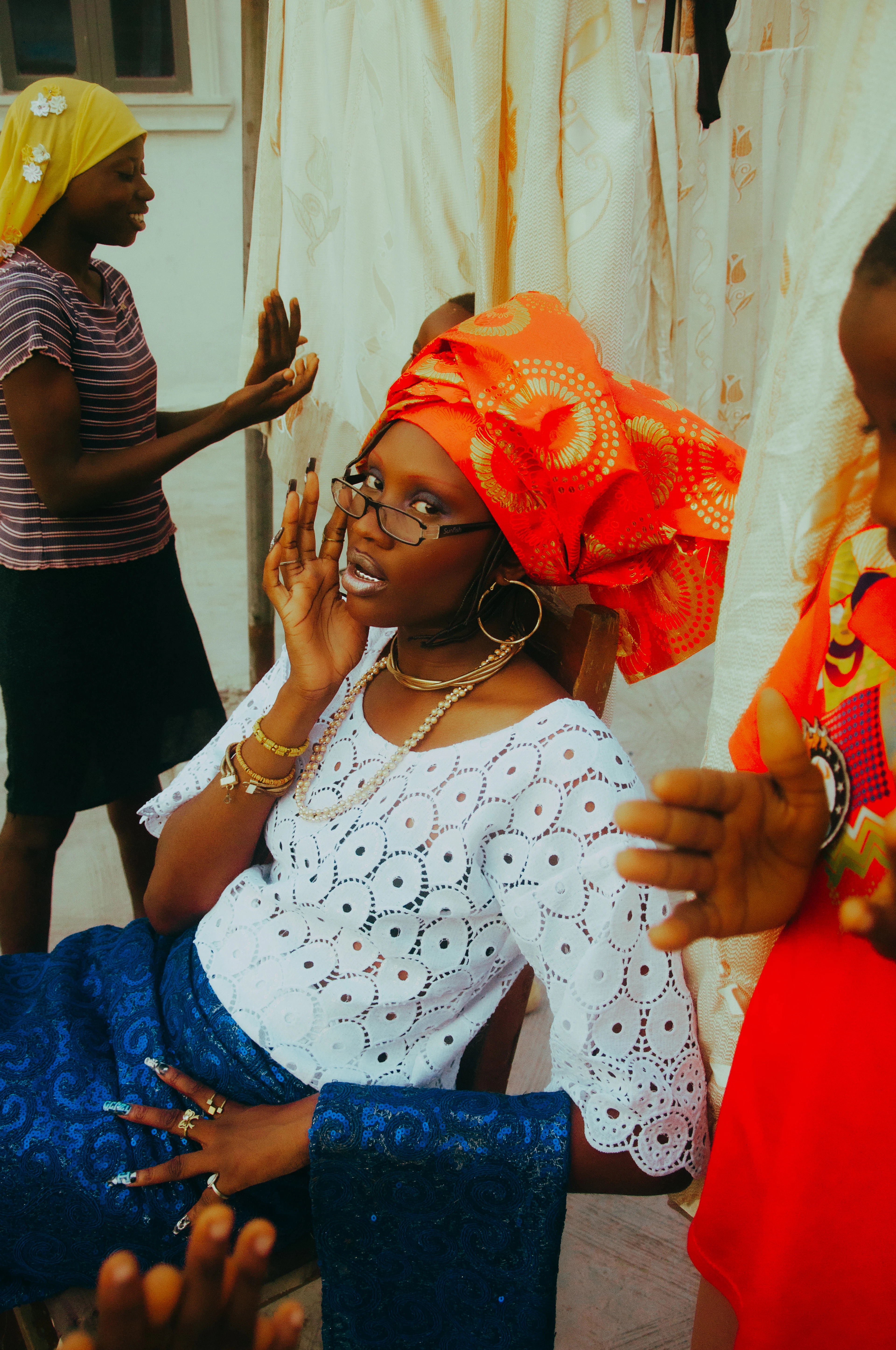 A woman in an orange head wrap and white lace top speaking, against a warm orange backdrop