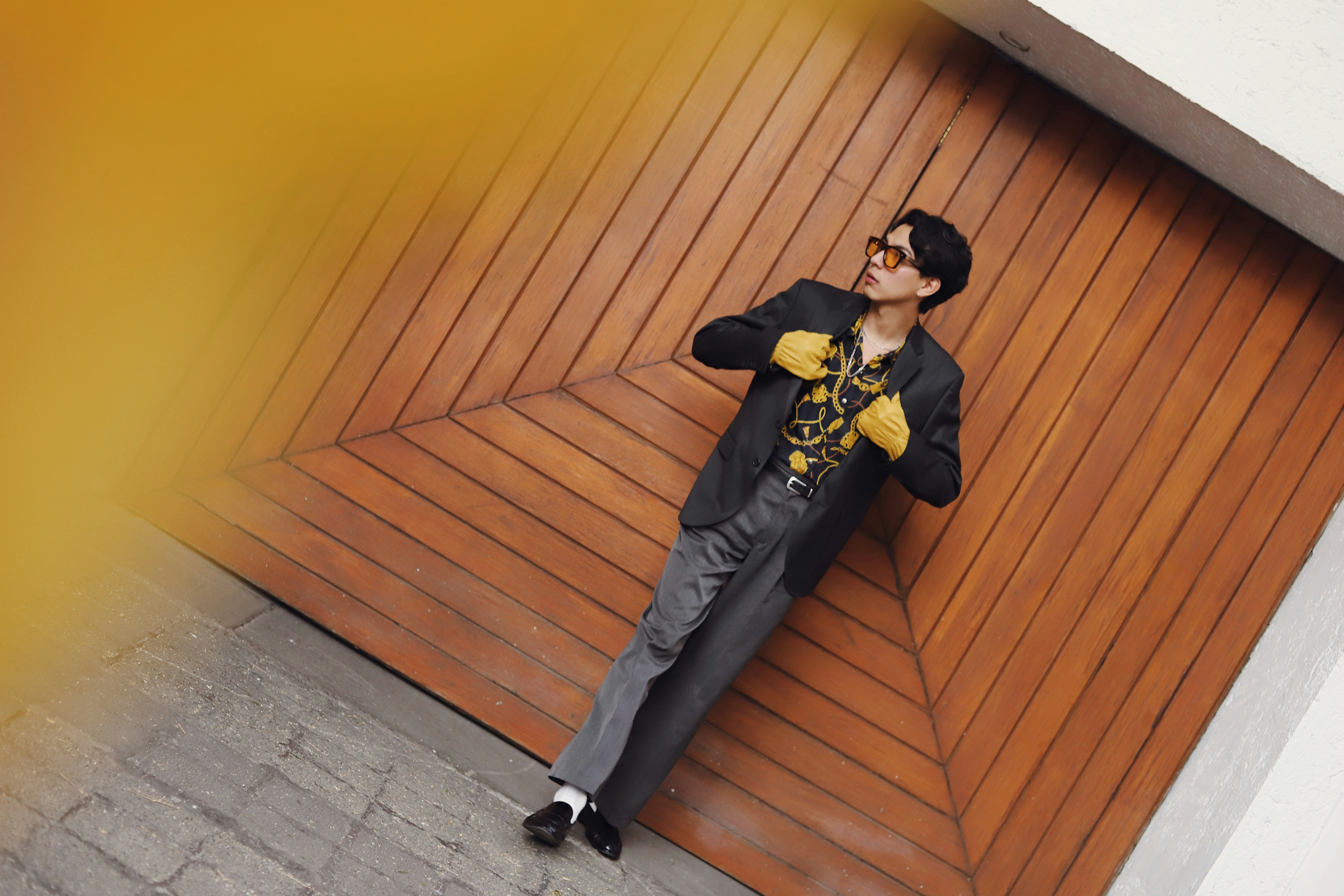 Man in stylish suit and yellow gloves leans against wall