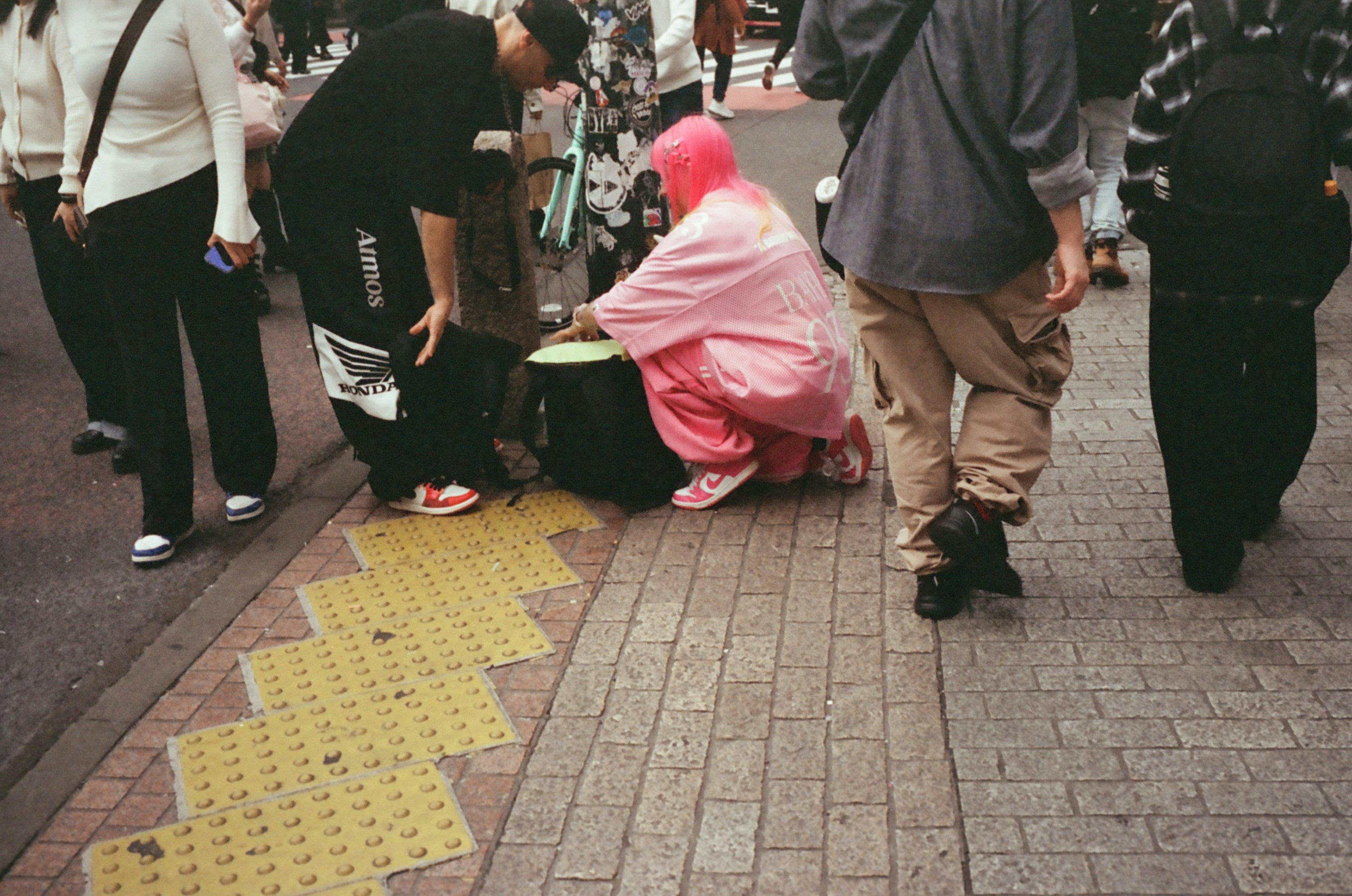 Three distraught women weeping near a covered manhole in an urban street, surrounded by a small crowd and police personnel. An ambulance in the background.
