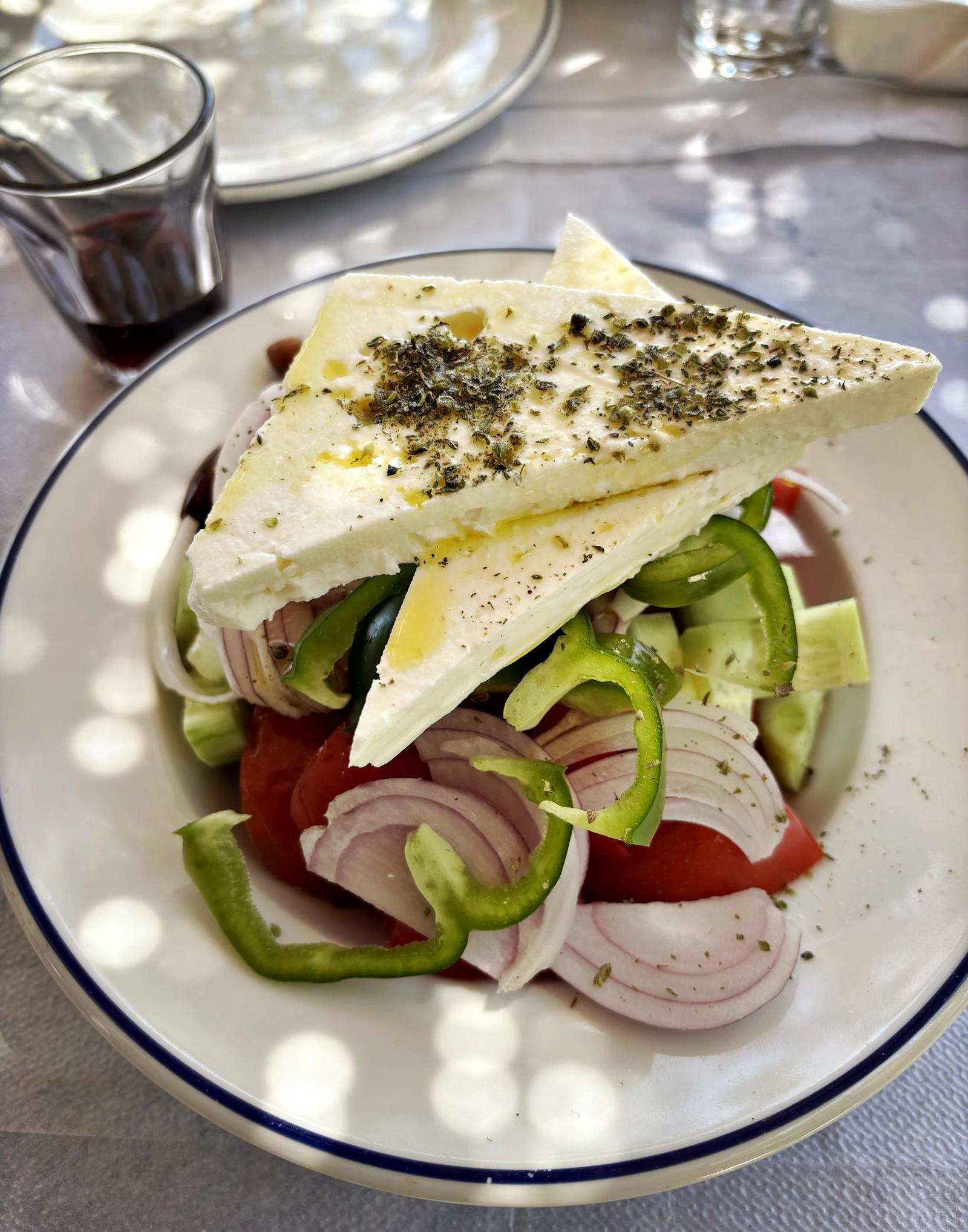 A classic Greek salad in a blue-rimmed taverna bowl, topped with a thick slab of feta drizzled with olive oil and oregano over tomatoes, red onion, green pepper and cucumber