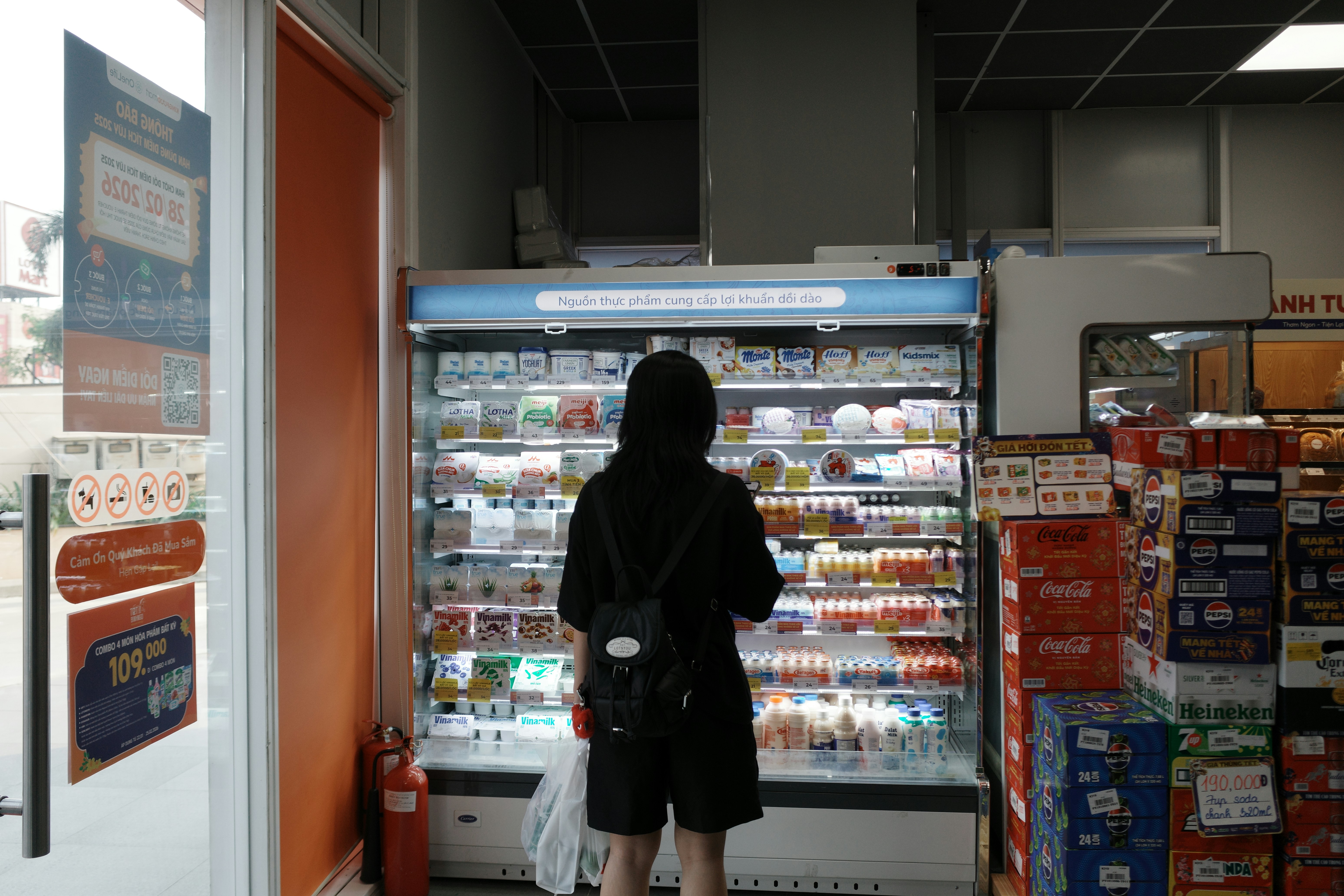 黒いパーカーを着た人がコンビニの冷蔵庫で店を買っている。