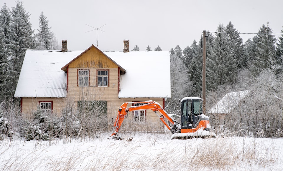 An orange compact excavator parked in deep snow beside a wood-sided two-story house ringed by snow-covered spruce trees on a gray Alaska winter day