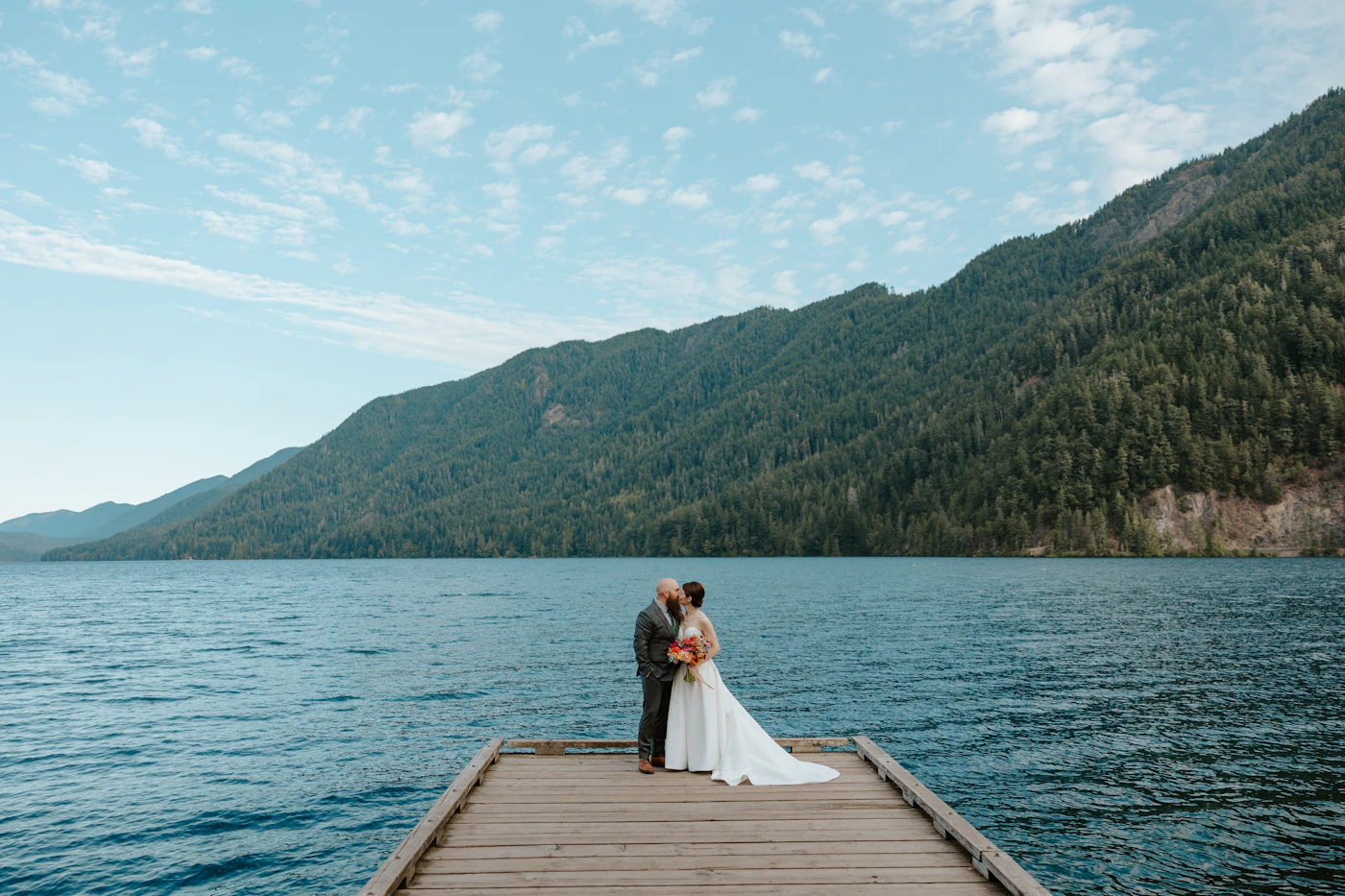 Couple standing close together on a wooden dock beside a calm reflective lake with mountains in the distance