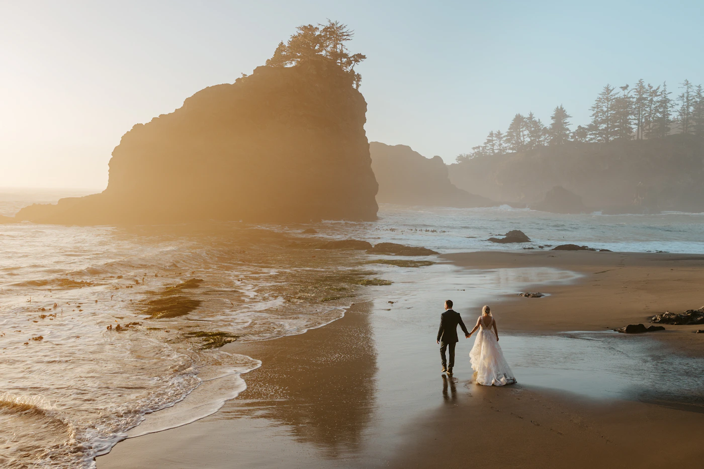 Couple in wedding attire walking together along an ocean shoreline at warm golden sunset