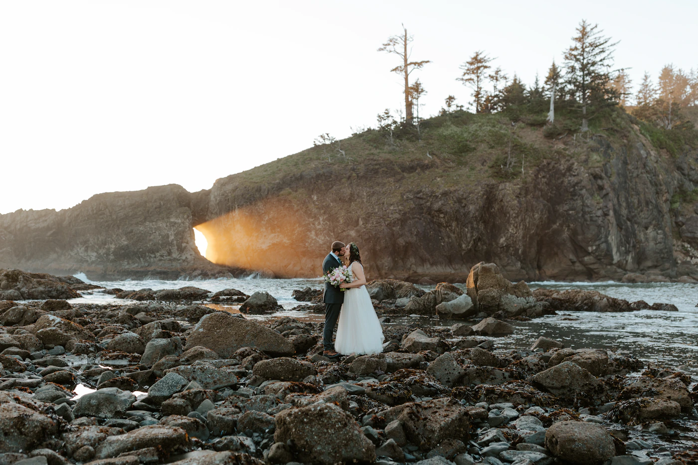 Newlywed couple in wedding attire on a rocky Pacific coastline at golden hour