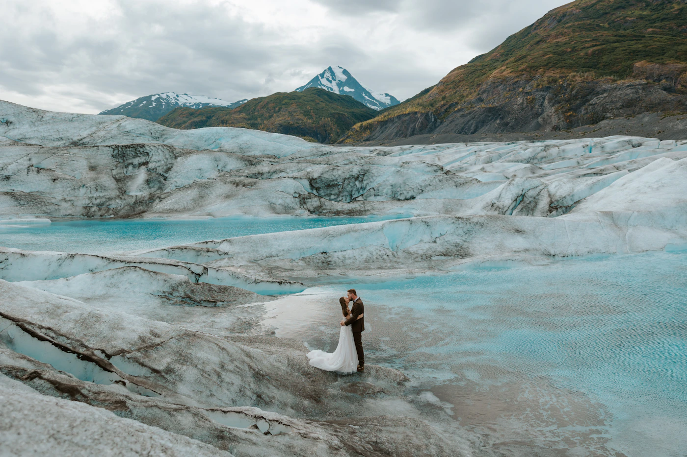 Couple in wedding attire standing together on a glacier with snow-capped mountain peaks rising behind them