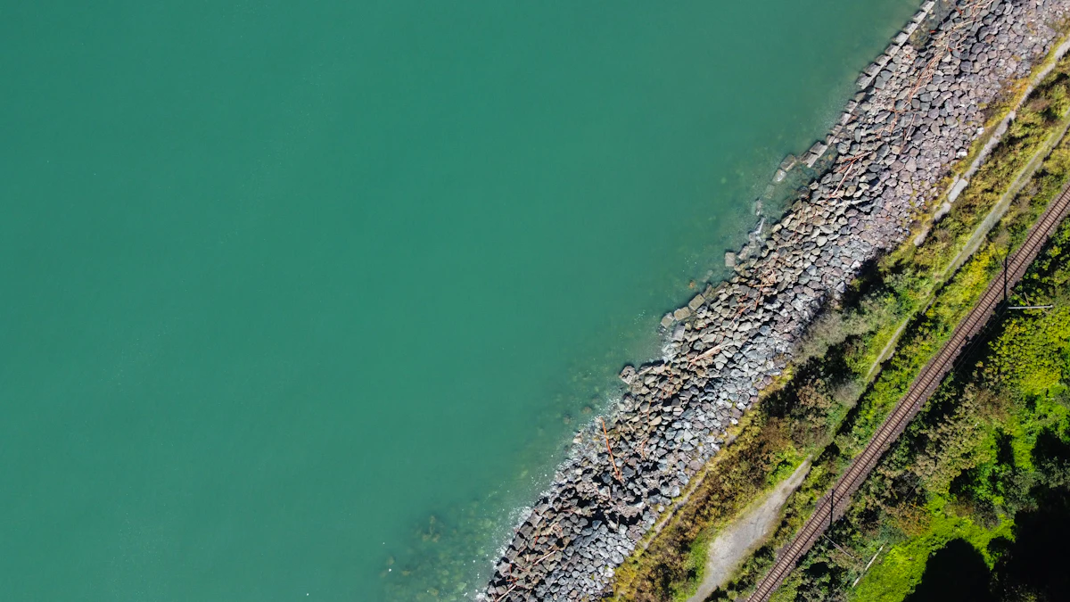 Aerial view of a rail line running along a rocky-toe embankment beside a body of water — the stream-parallel reach where Texas Tuff Rock Bags and Rock Logs hold the toe against runoff and channel flow