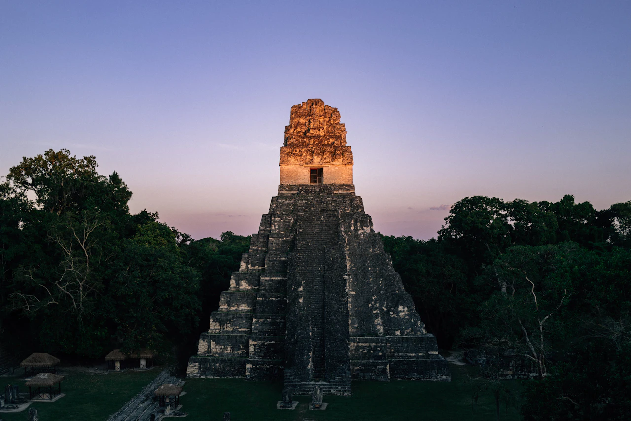A steep stone temple pyramid rising above the forest canopy, its top lit orange by the last light of the day.