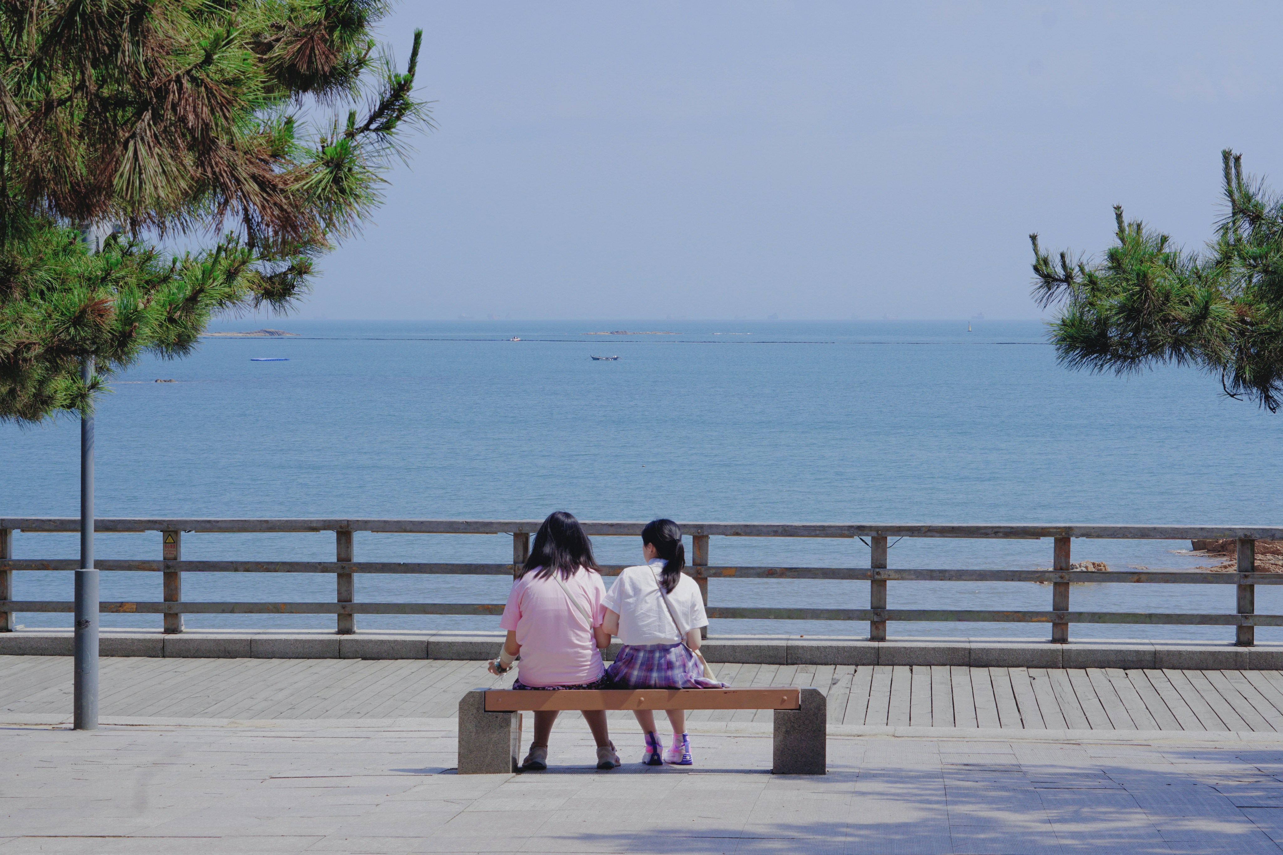 Two girls sit on a bench overlooking the ocean.