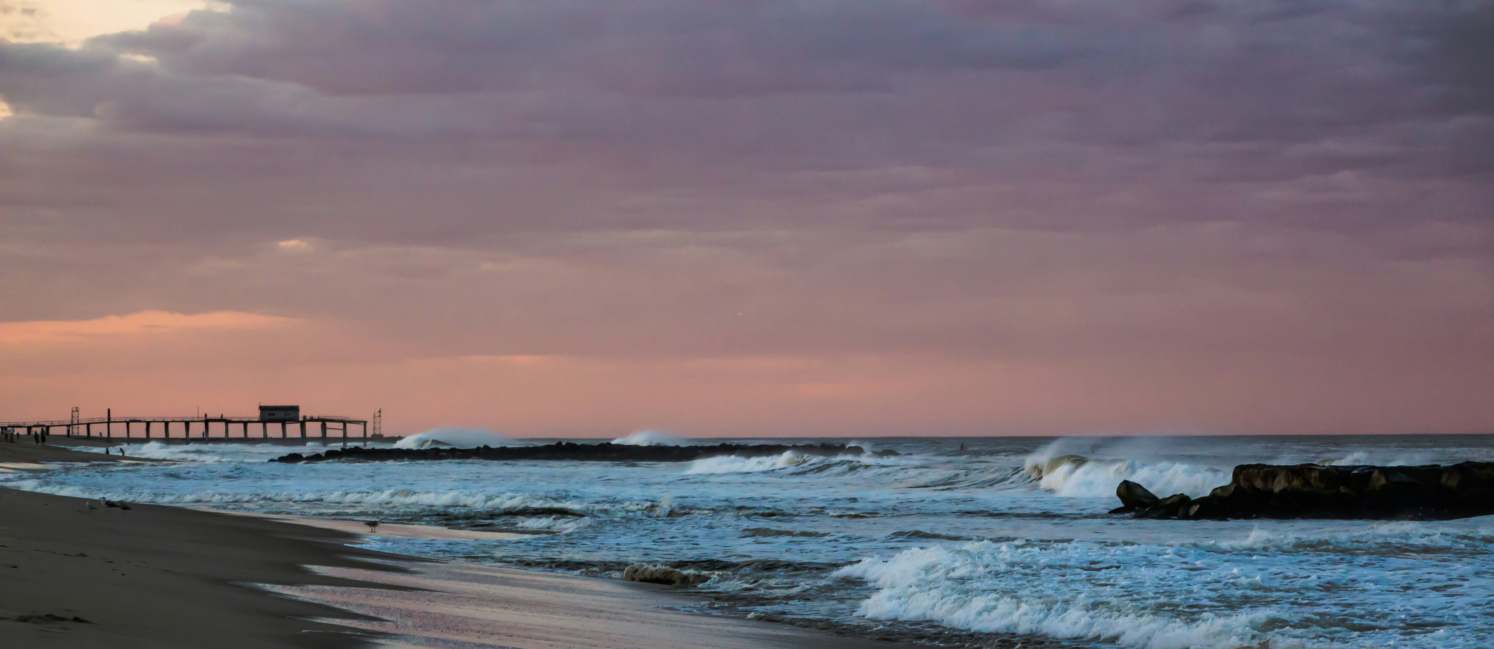 Waves crash on a beach near a pier at sunset.