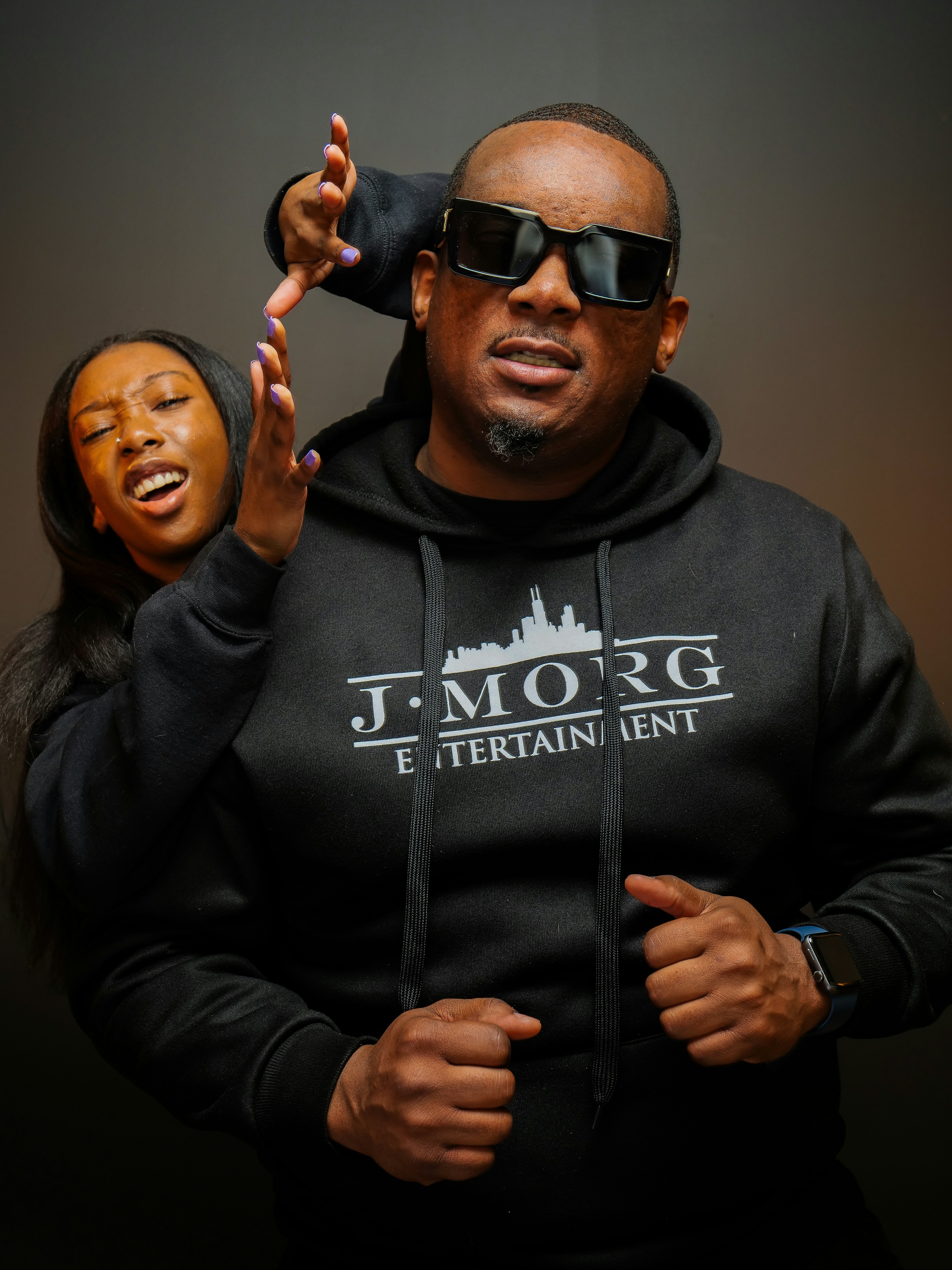 Man in sunglasses and hoodie with woman behind him.