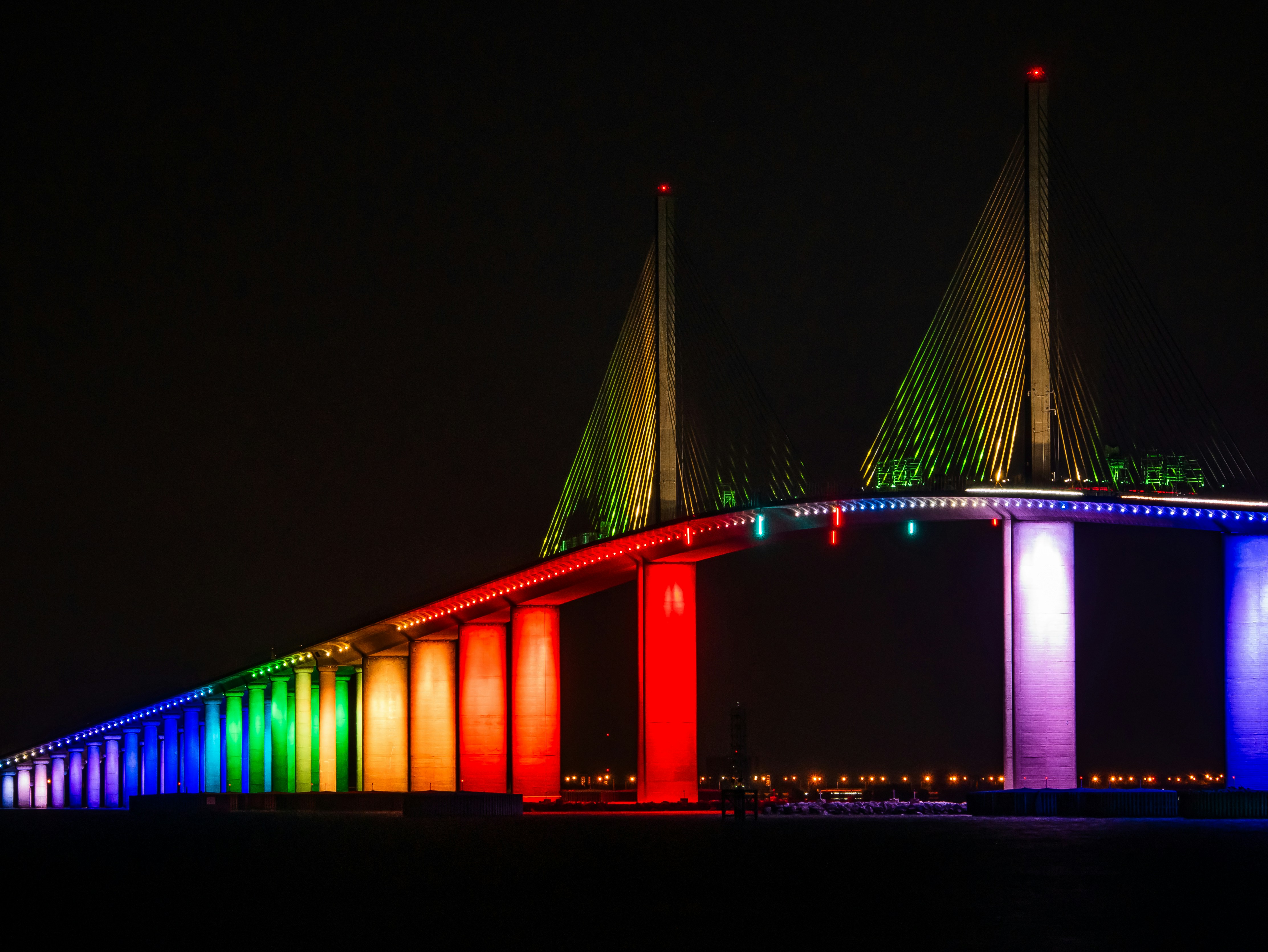A cable-stayed bridge illuminated with rainbow lights at night.