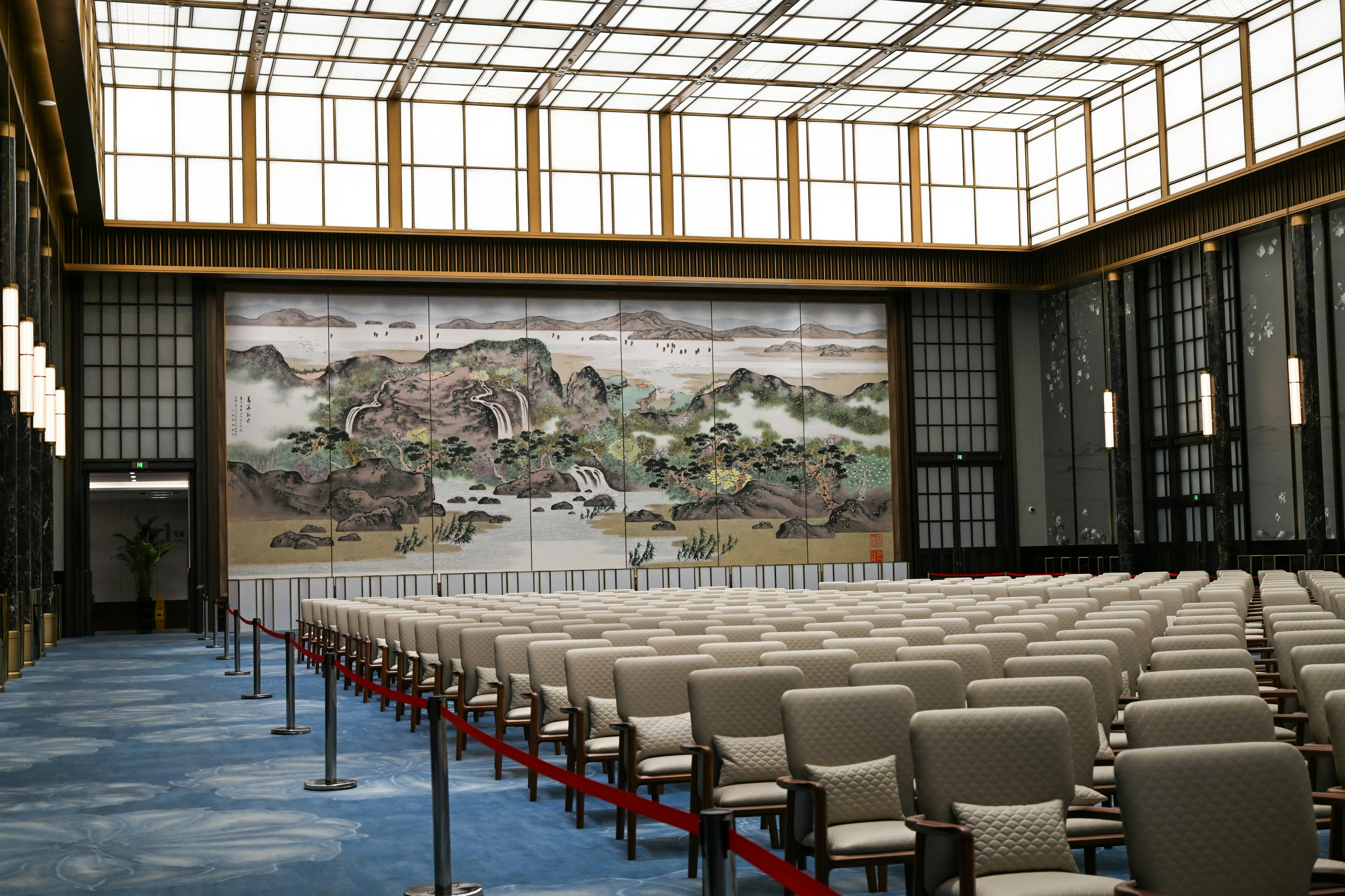 A panoramic shot of a modern, well-lit conference hall bustling with attendees and speakers on stage at an AI summit, with large screens displaying AI-related graphics.