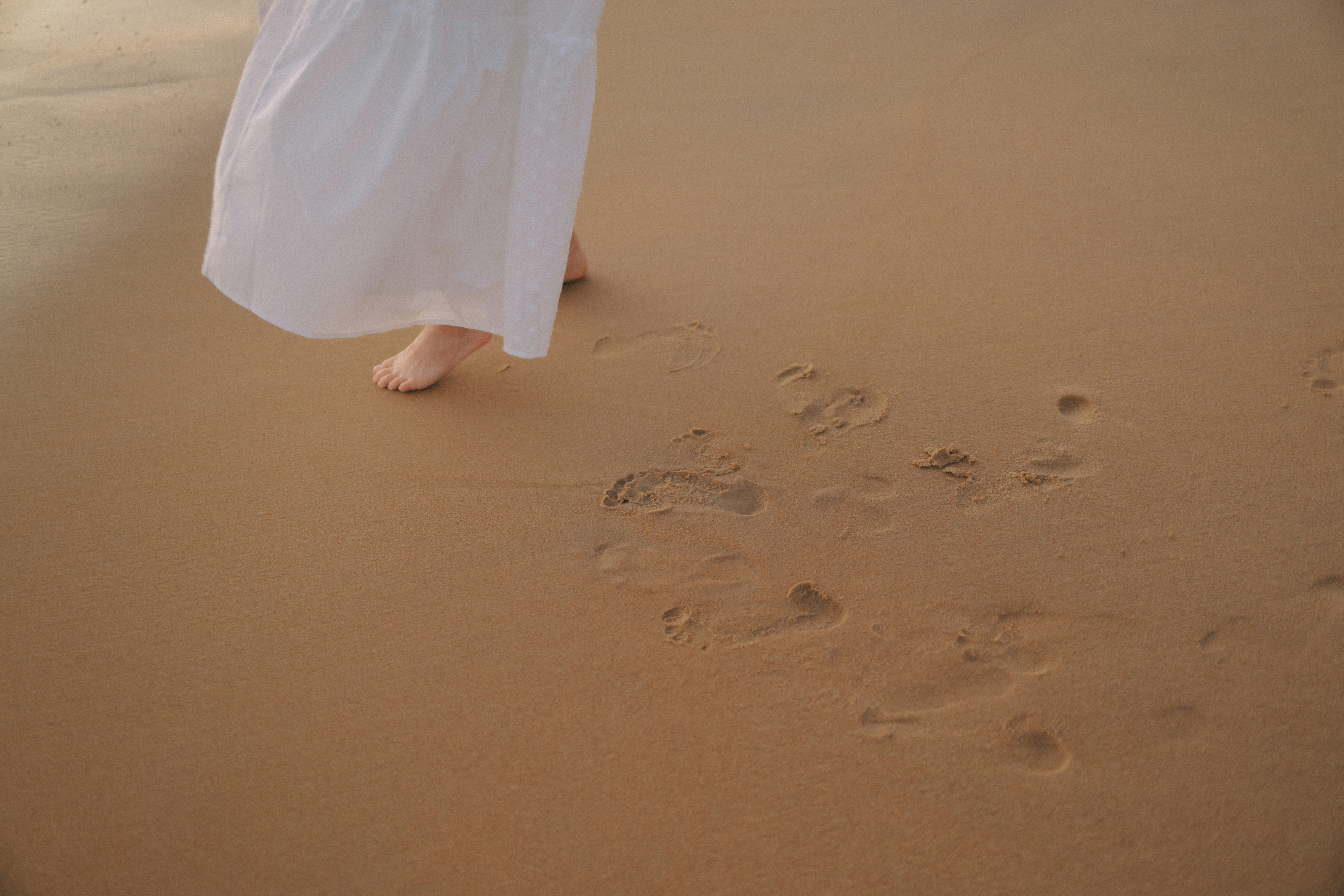 Bare feet walking on a sandy beach leaving footprints.