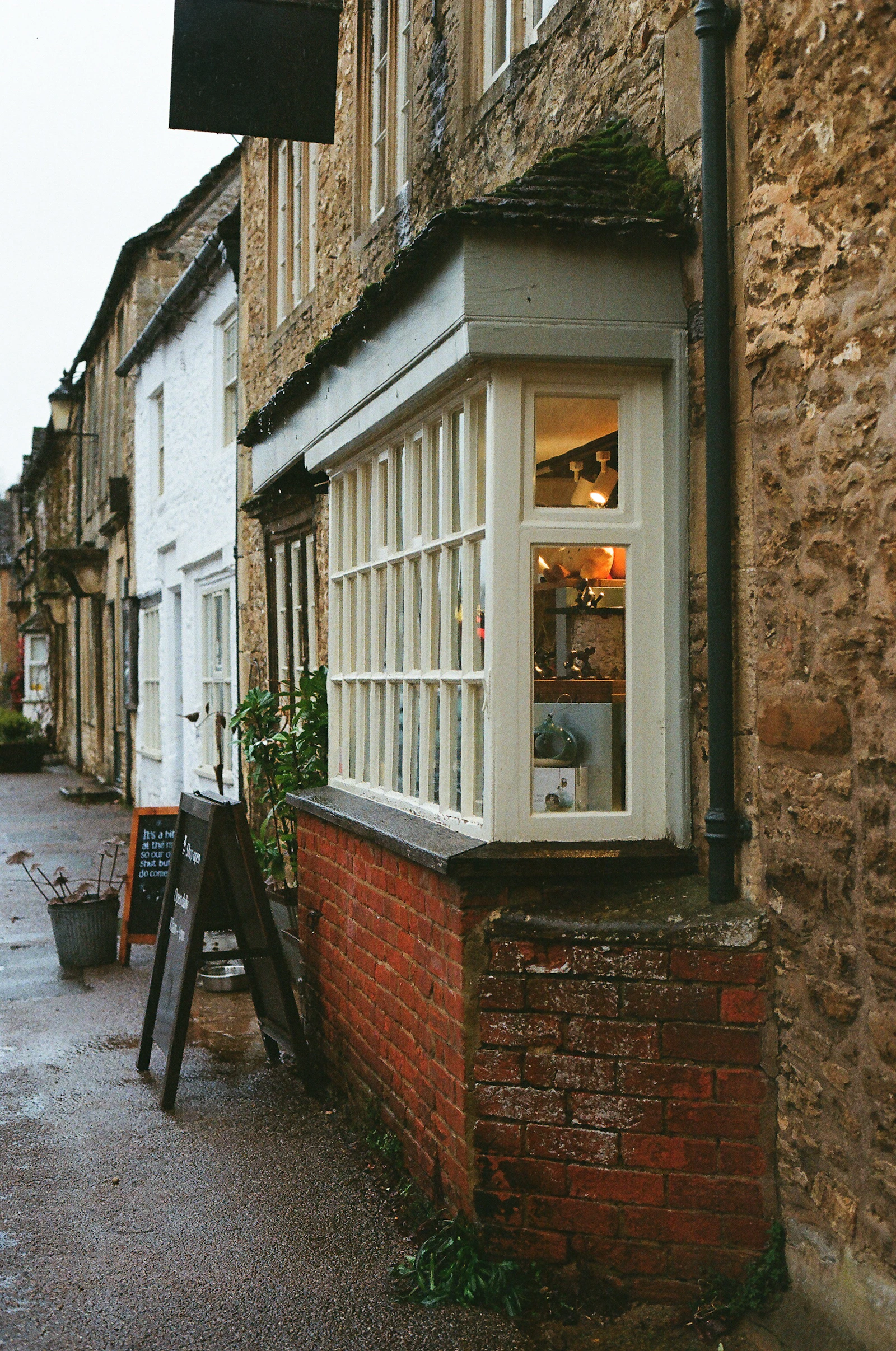 A shop's white-framed bay window jutting out over a red brick plinth on a wet cobbled street at dusk, the room inside lit warm and a chalkboard propped against the wall beside it.