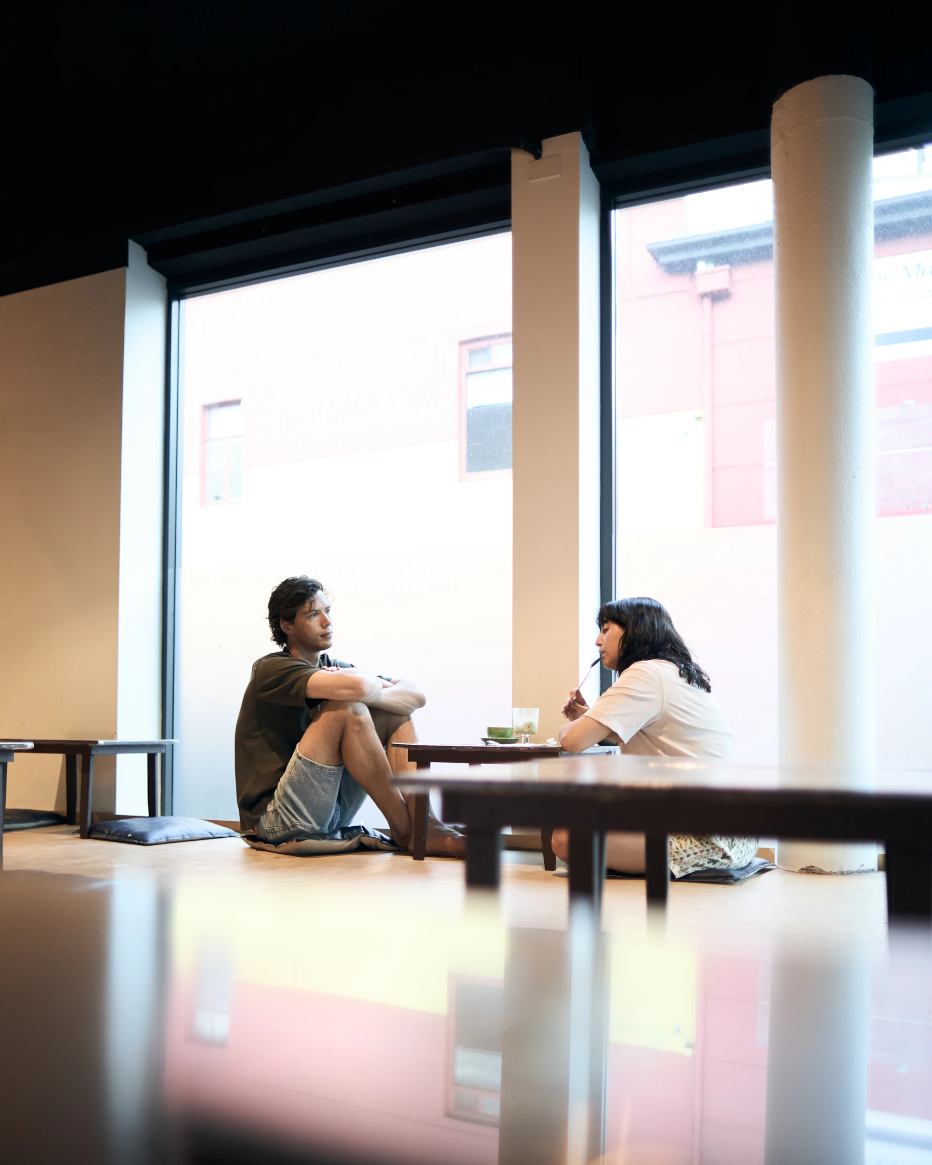 Two people sitting on floor talking near large window