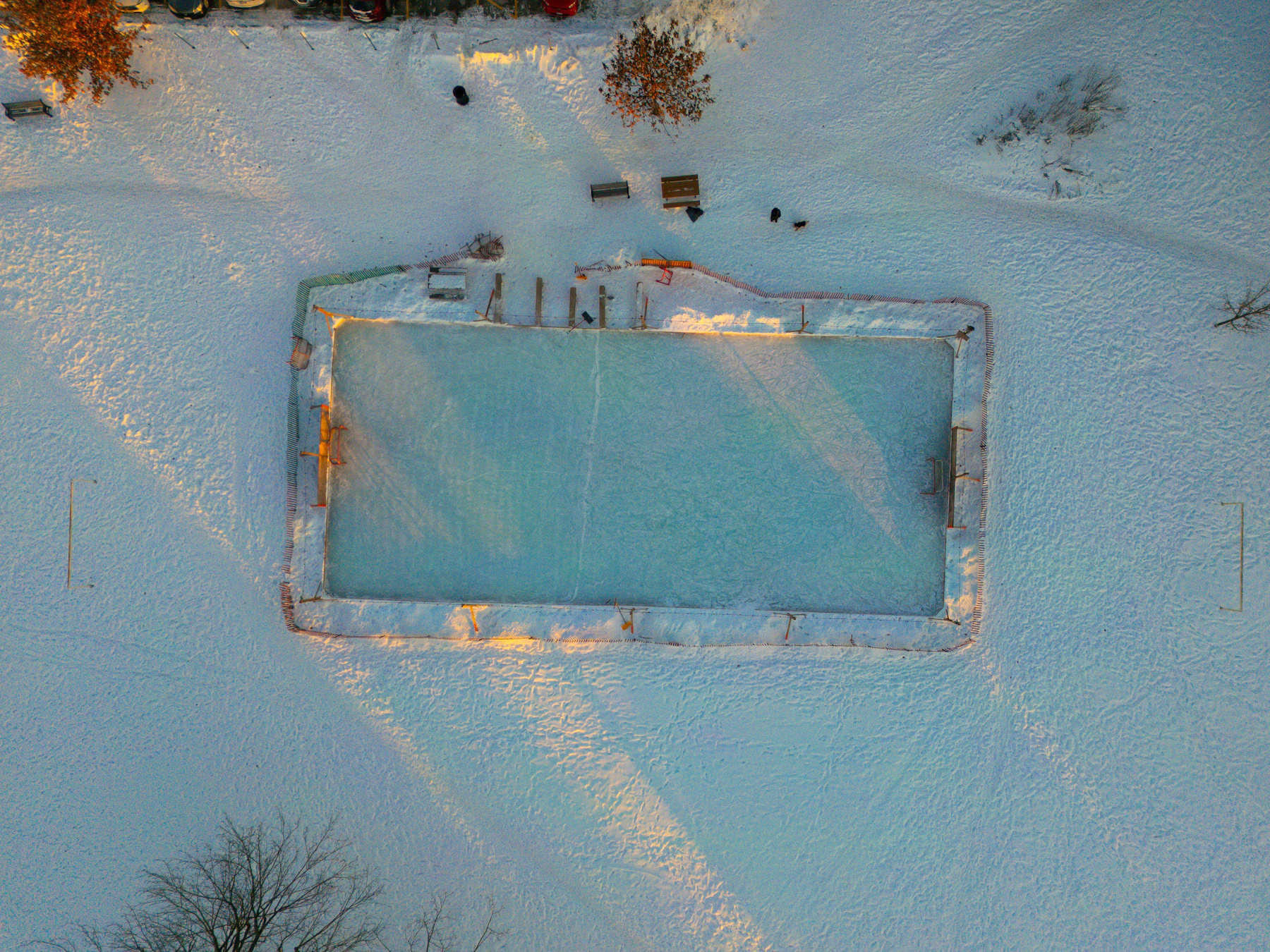 A small rectangular ice rink framed by boards and snowbanks in a snowy yard, seen from directly above