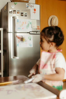 Young child in apron near refrigerator with drawings.
