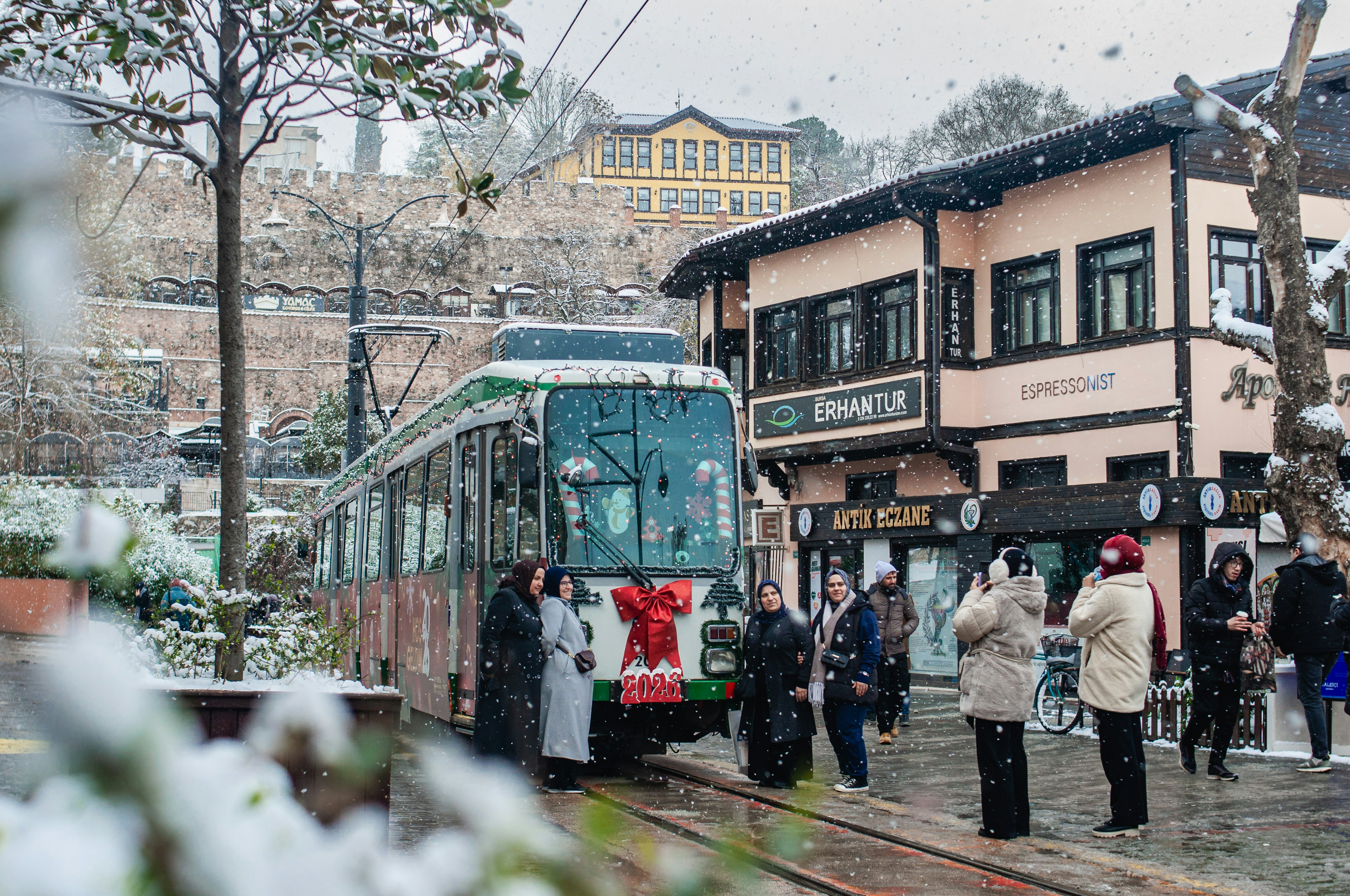 People wait for tram on snowy street with buildings.