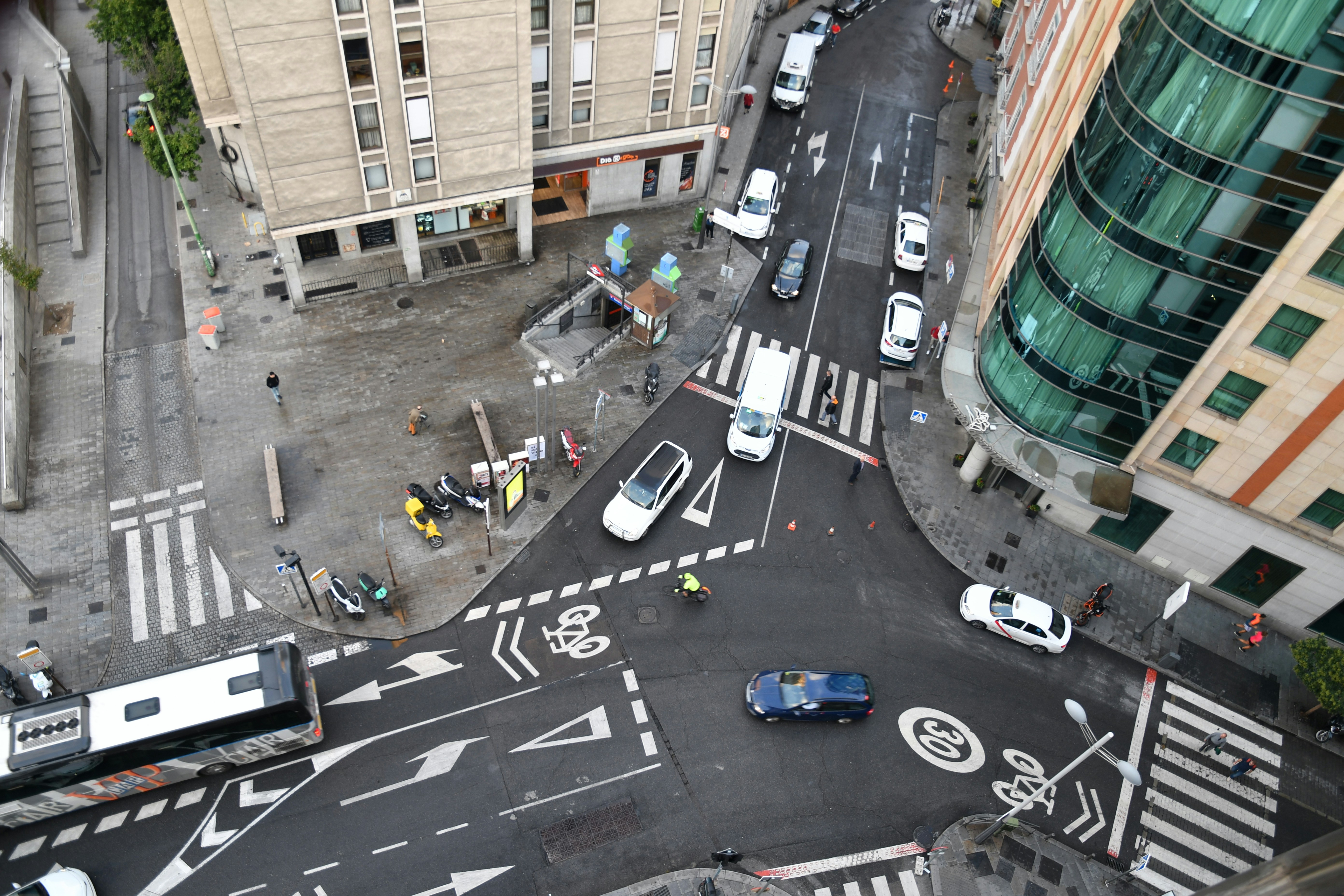 An aerial view of a street, with cars and motorcycles moving through lanes, intersections, and surrounding buildings in downtown Madrid, Spain.