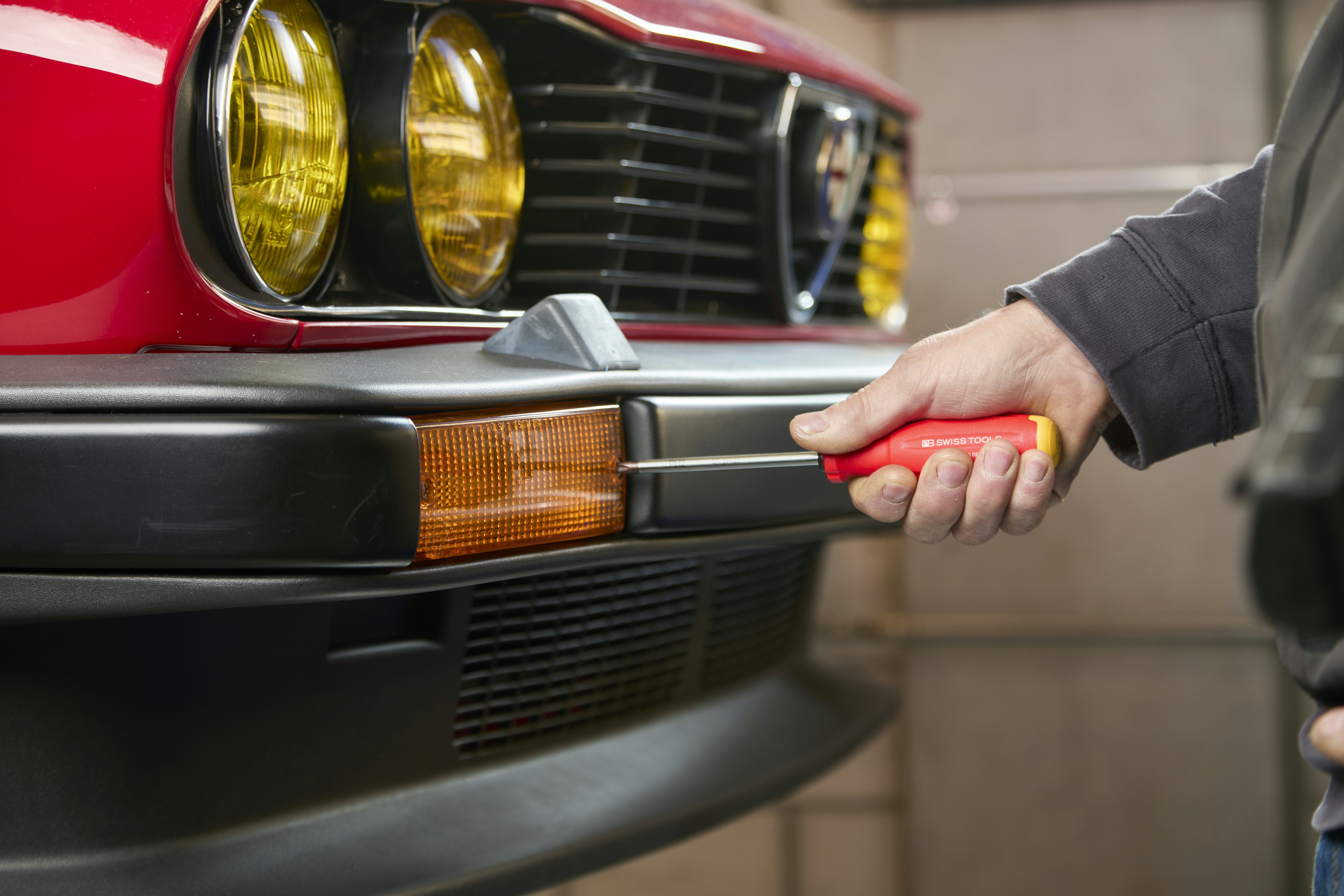 Mechanic repairs a red car's bumper with a screwdriver.