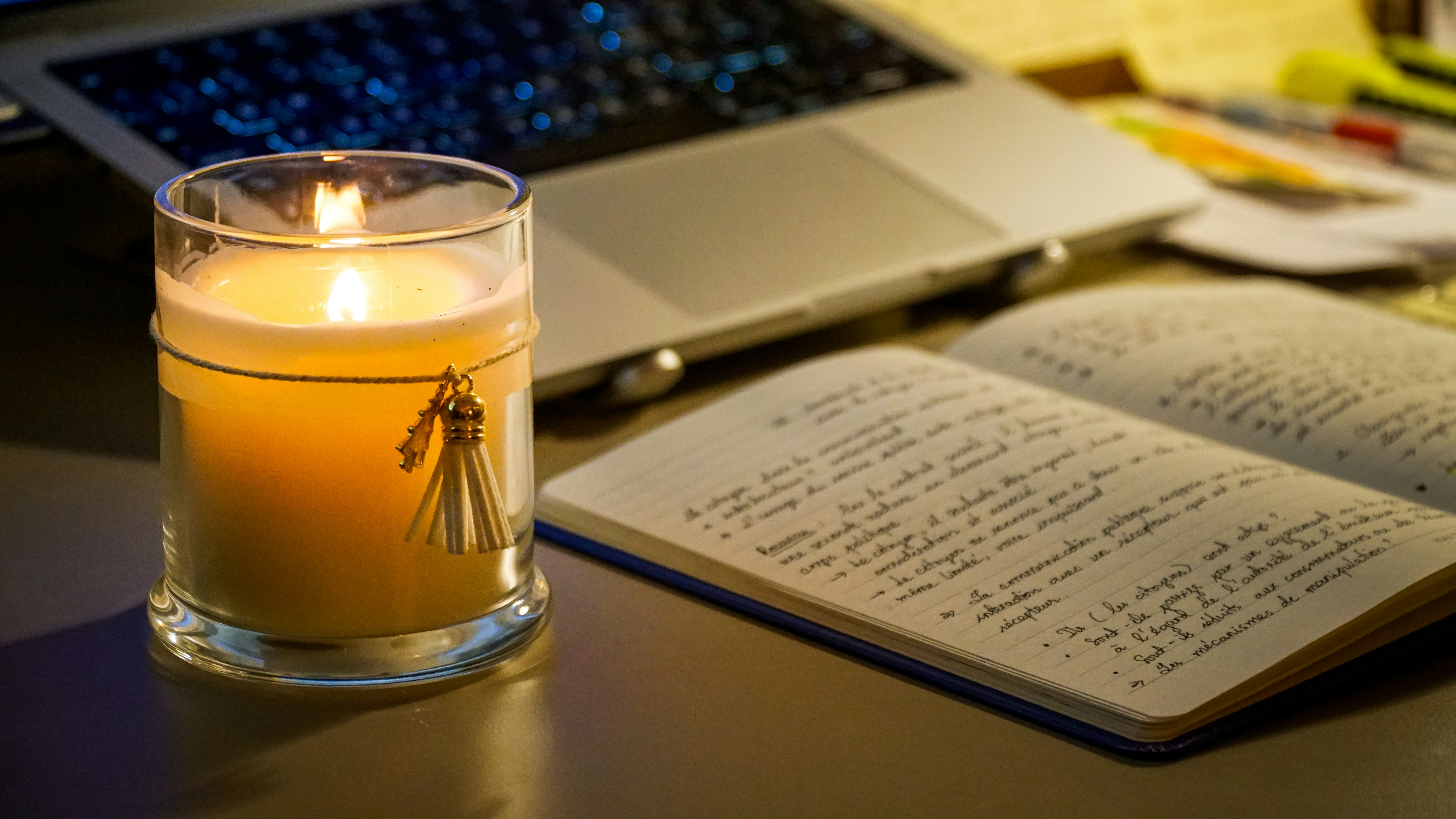 A lit candle next to an open notebook and laptop.