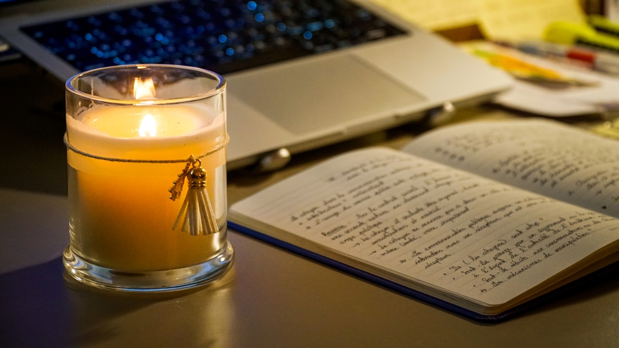 A lit candle next to an open notebook and laptop.