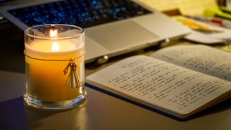 A lit candle next to an open notebook and laptop.