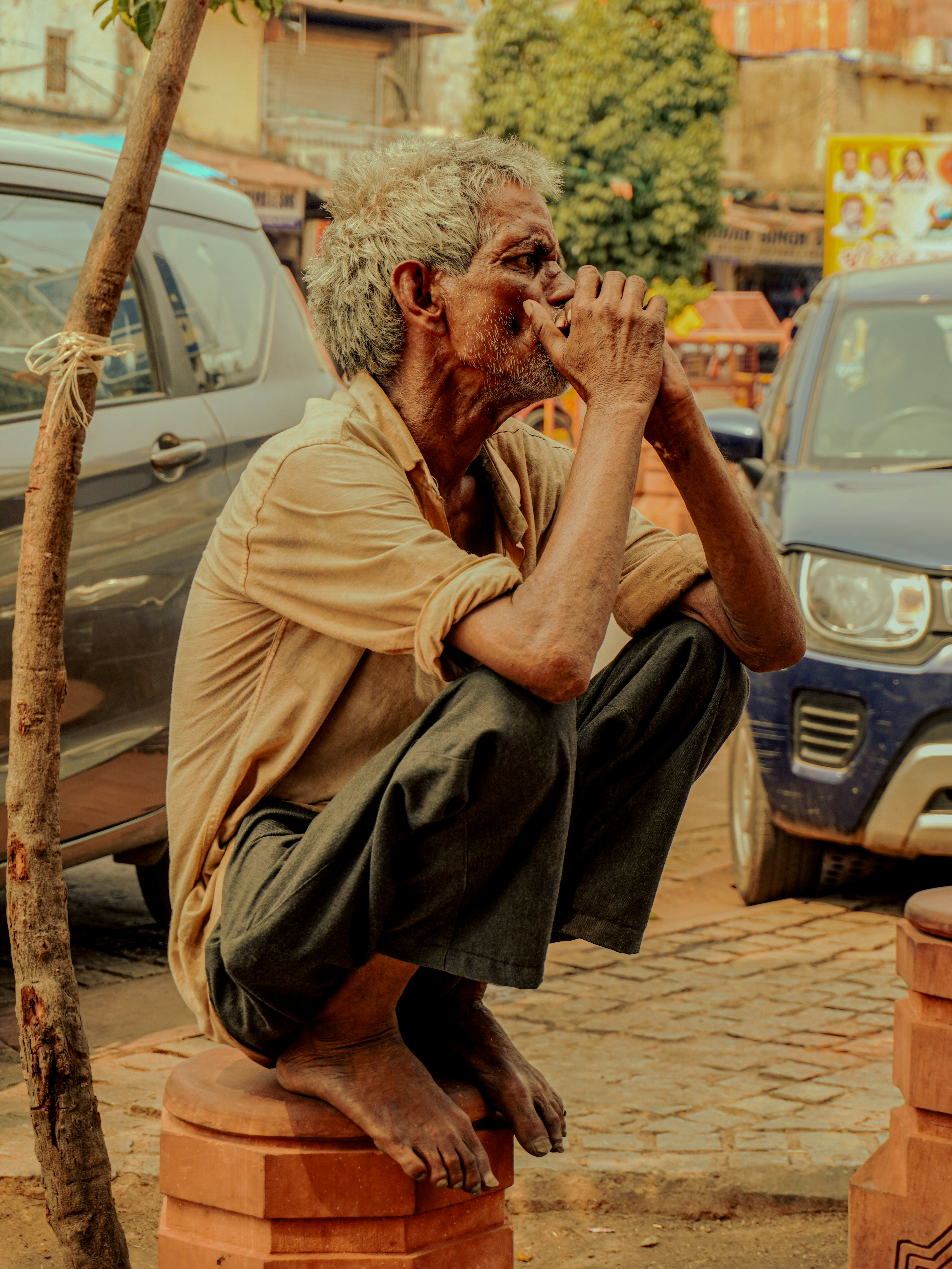 Elderly man squats with hands clasped near face.