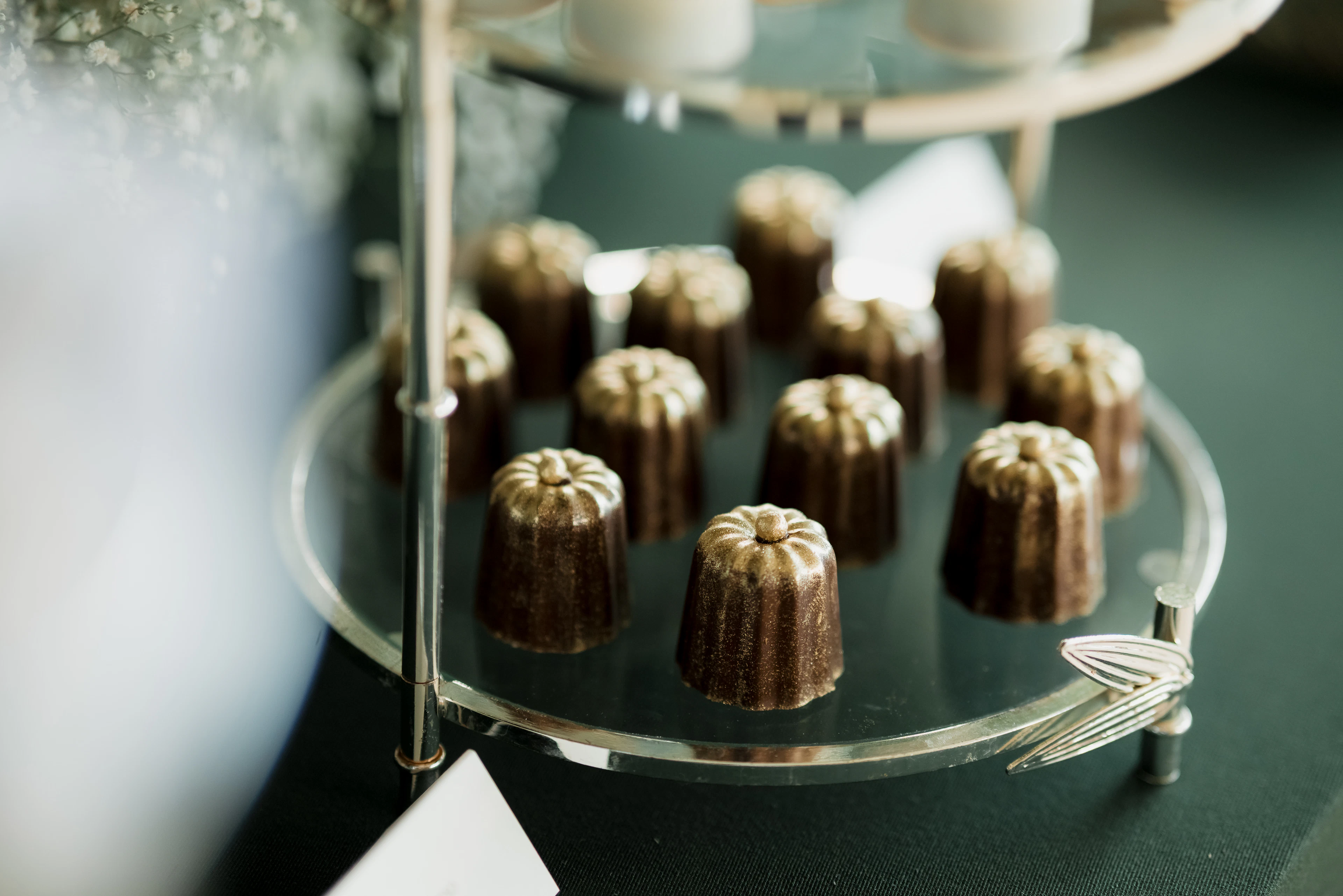 A dozen canelés de Bordeaux, small fluted pastries with dark glossy caramelised crusts, arranged in rows on a glass tiered stand against a deep green cloth.