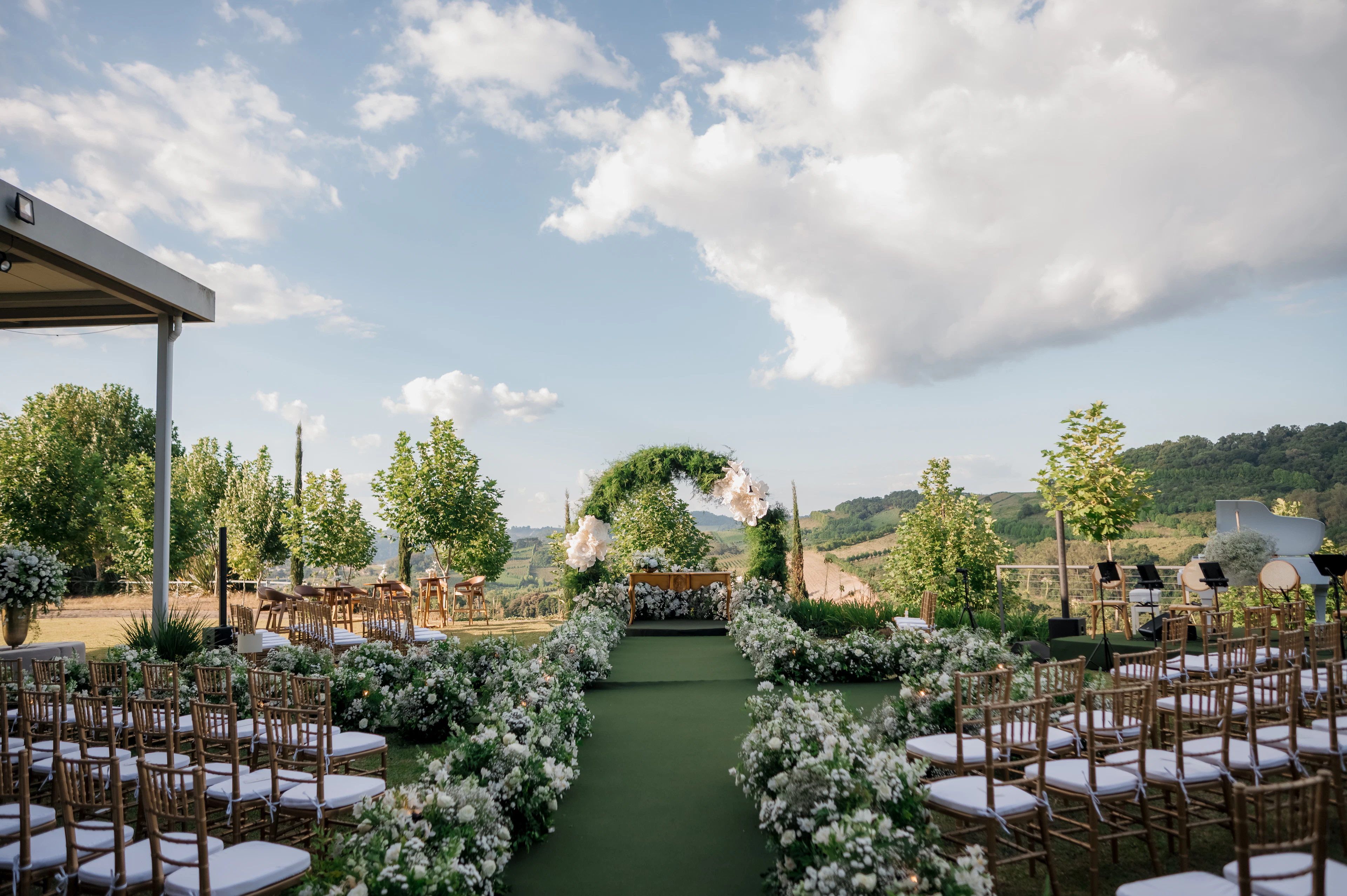 An outdoor hillside ceremony: rows of chairs along a green aisle of white flowers leading to a floral arch.