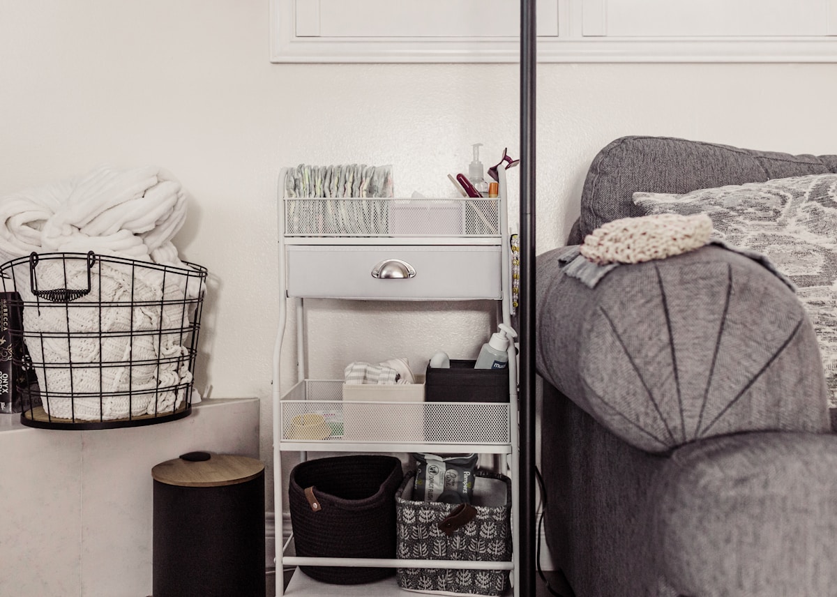 White rolling three-tier metal storage cart with mesh baskets full of organizers beside sofa with wire basket holding folded blankets