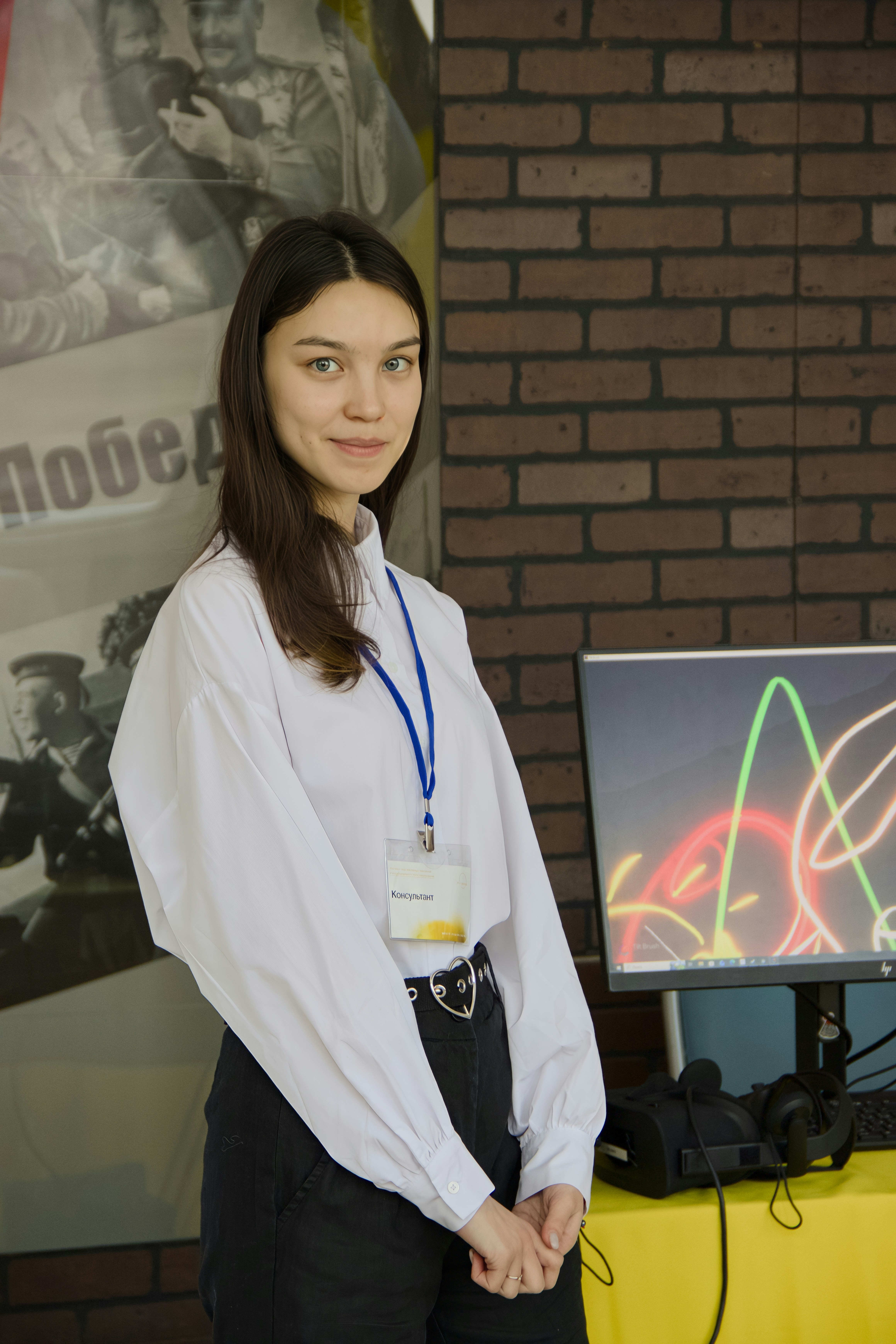 Young woman in white shirt with lanyard stands near computer.