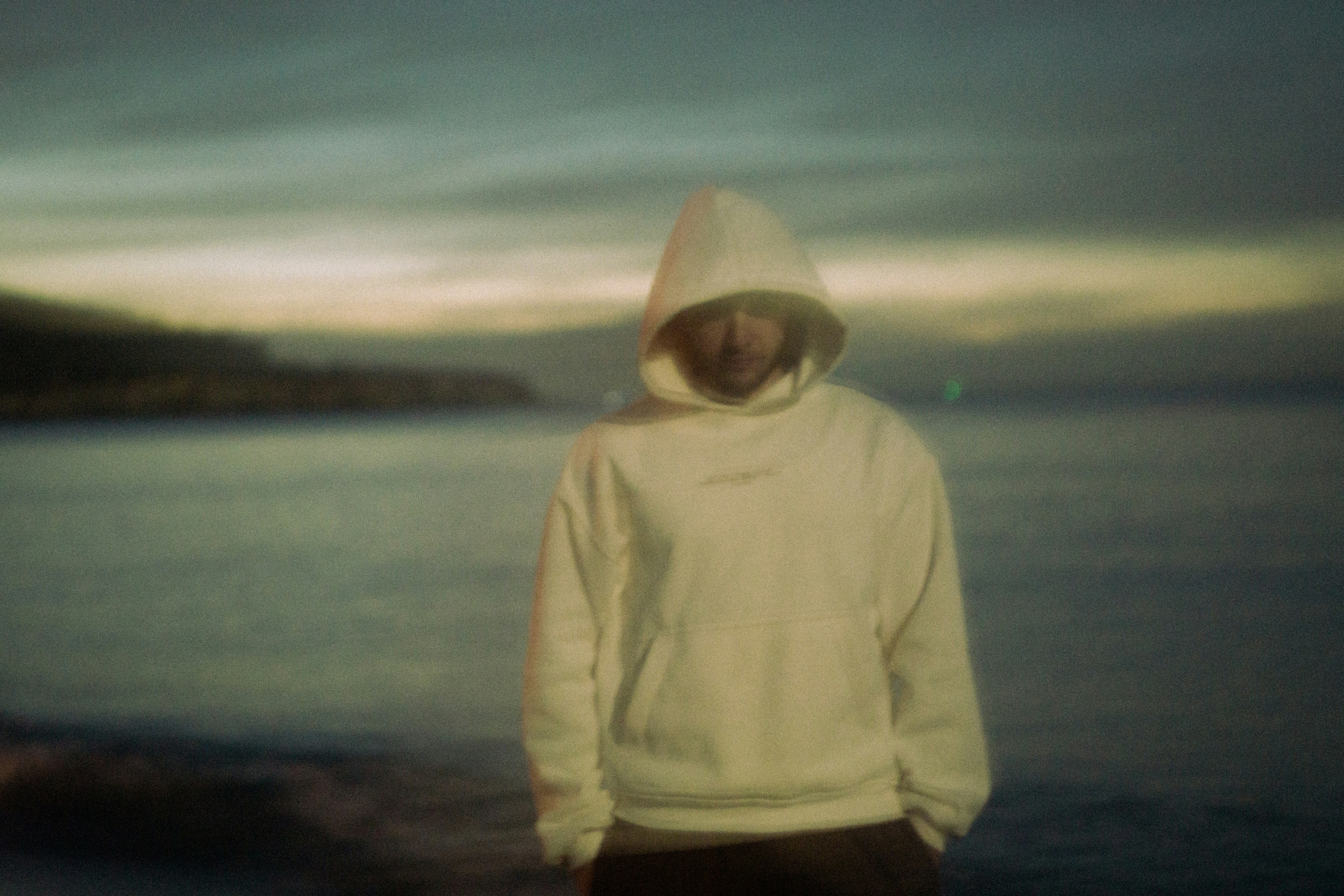 Person in hoodie stands by the ocean at dusk.