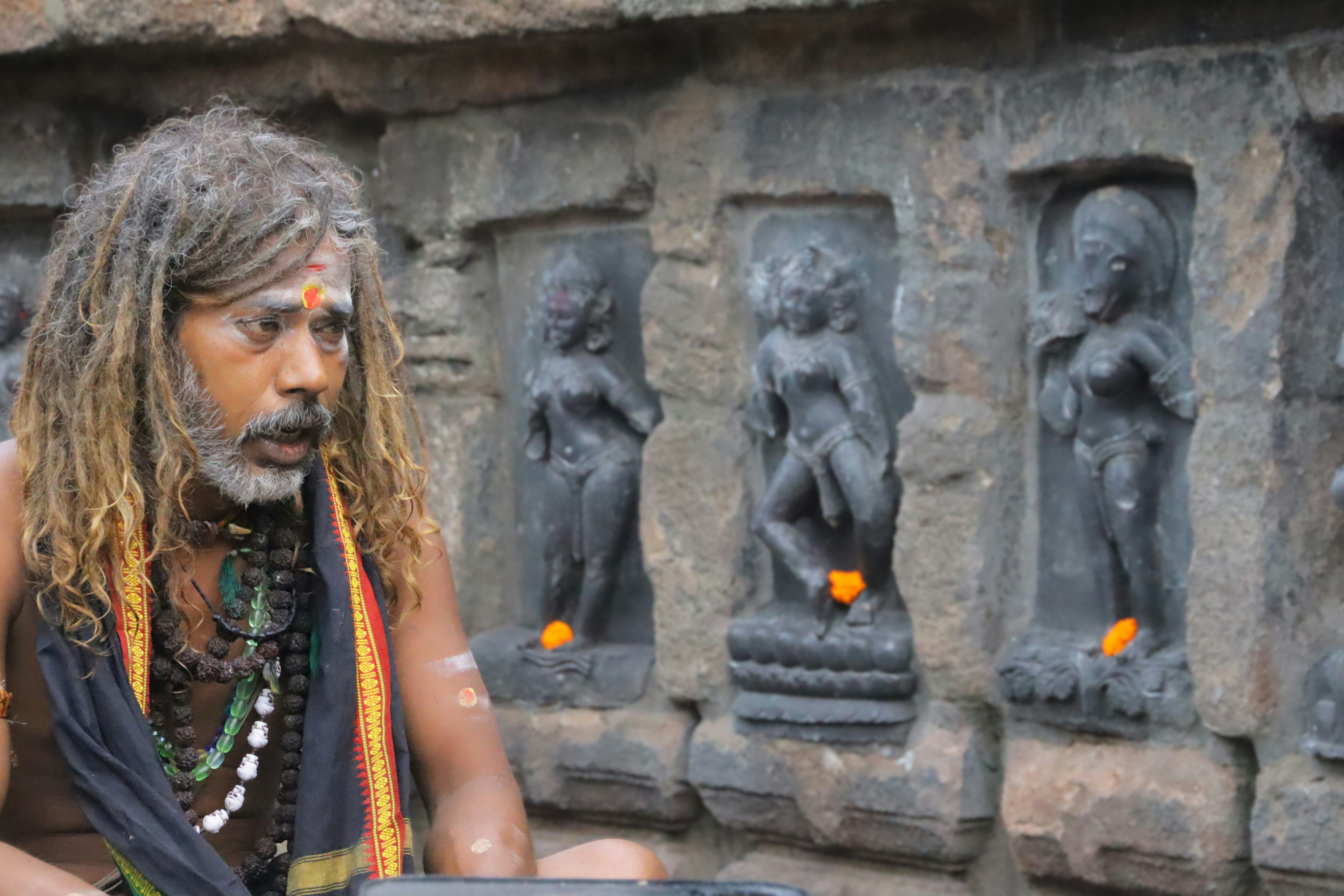 Man with dreadlocks and religious markings sits near stone carvings.