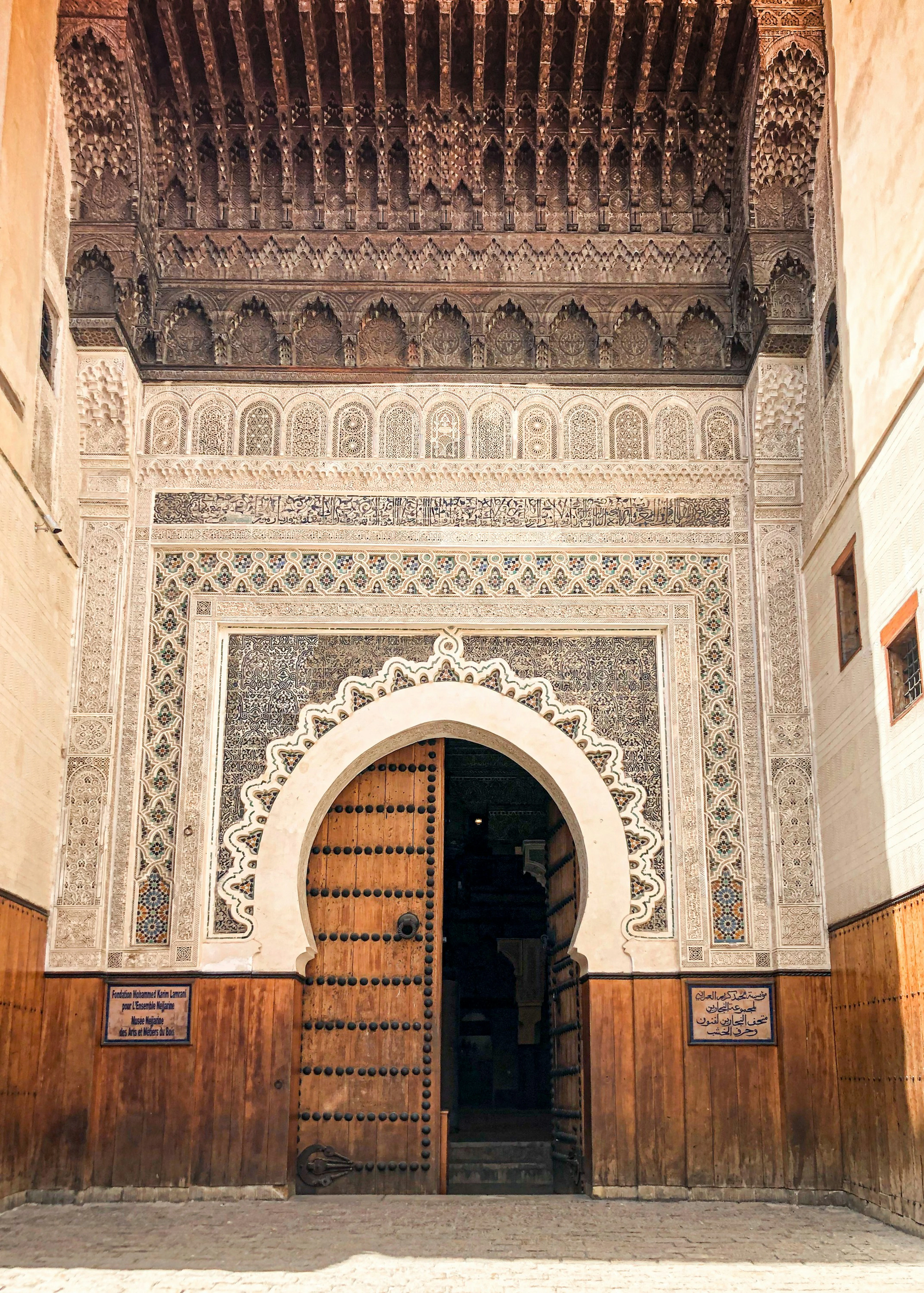 The carved cedar canopy, stucco and zellij portal of the Nejjarine Museum of Wood Arts on Nejjarine Square in the Fez medina, its heavy studded door standing open.