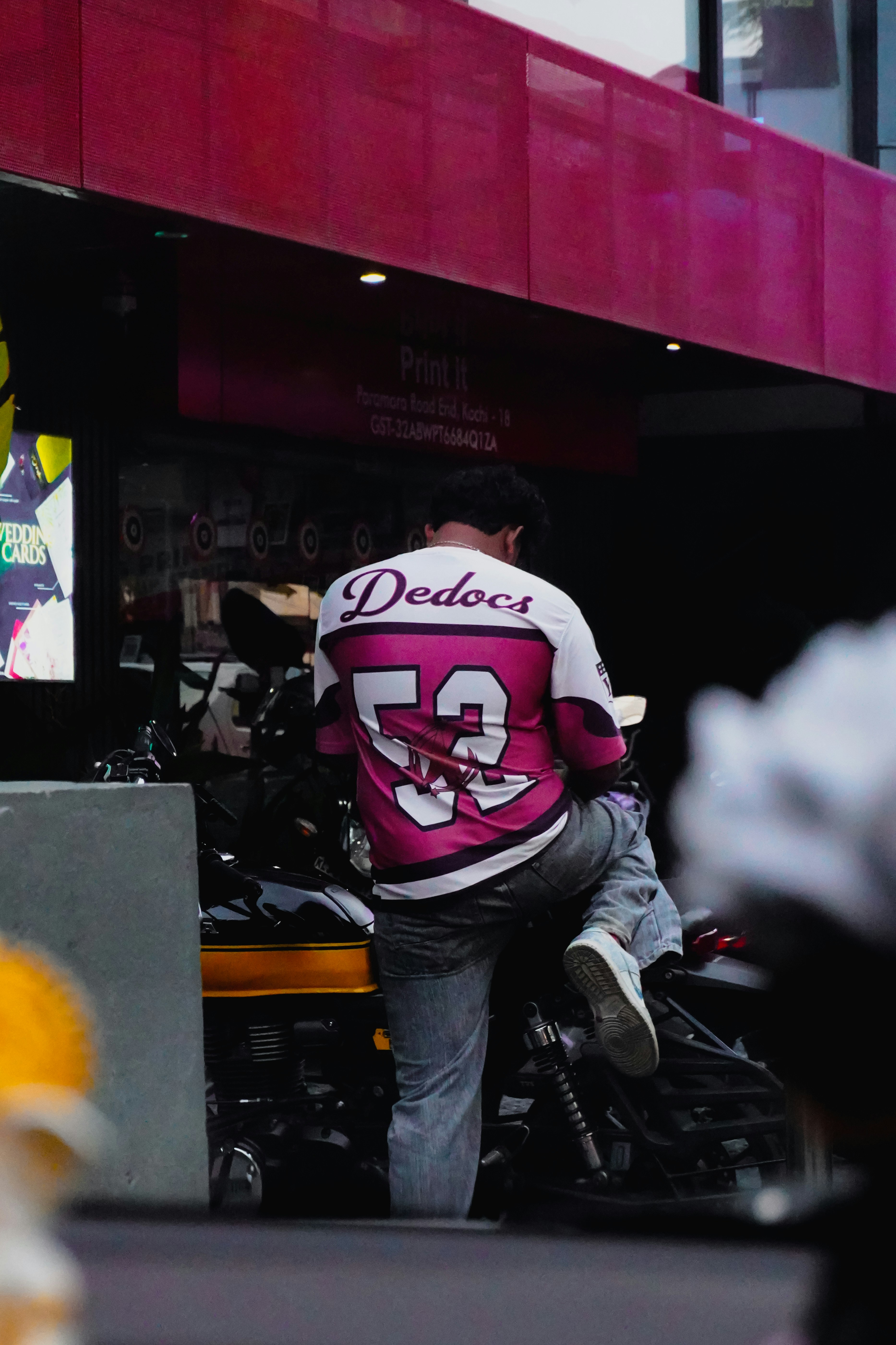 Man in pink and white jersey sits on motorcycle.