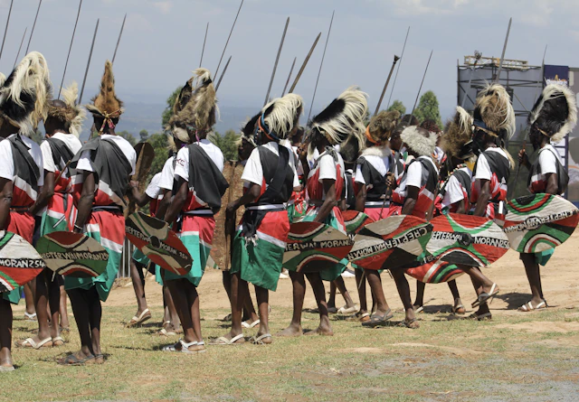 A line of Maasai warriors in colorful traditional attire, holding spears and painted shields, standing outdoors during a ceremony in Nandi, Kenya.